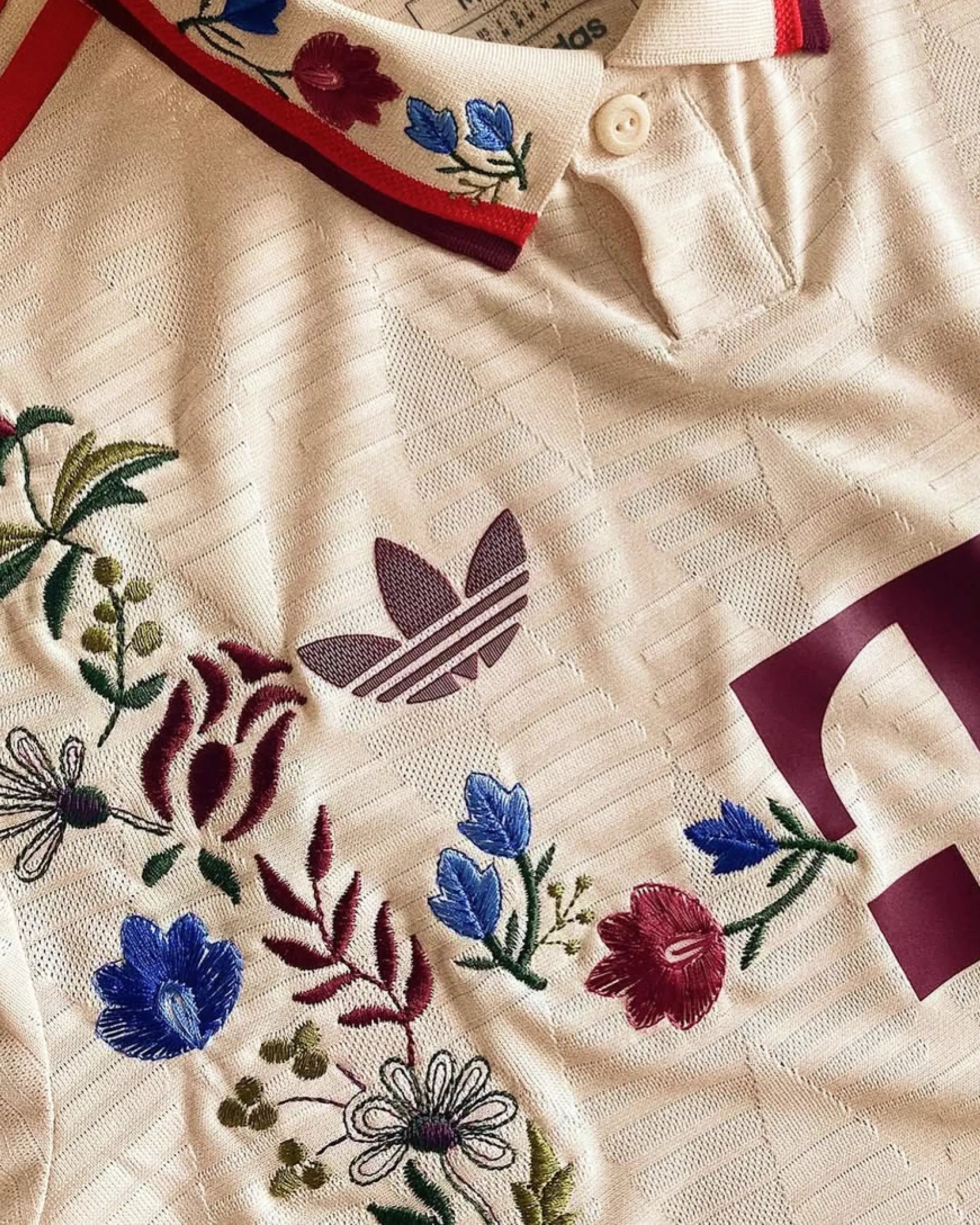 Close-up hand-embroidered Bavarian folk flowers — burgundy botanicals, blue campanula, white daisies and olive foliage on Bayern Munich cream concept jersey — artisanal embroidery VOLTA Paris & Versailles