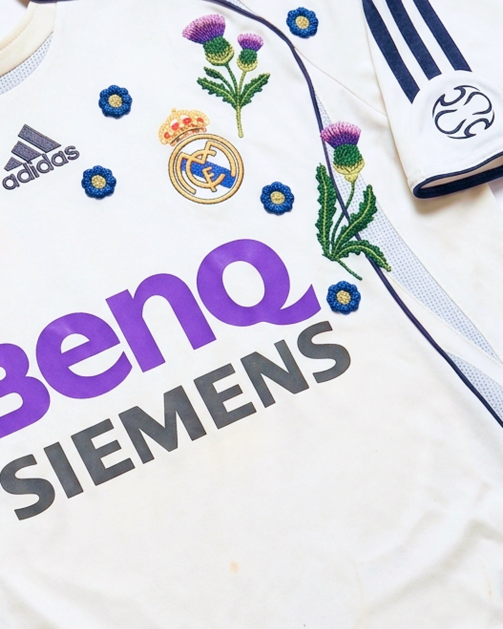 Close-up hand-embroidered purple Scottish thistle and blue botanical flowers around Real Madrid crest on retro concept jersey — artisanal embroidery VOLTA Paris & Versailles