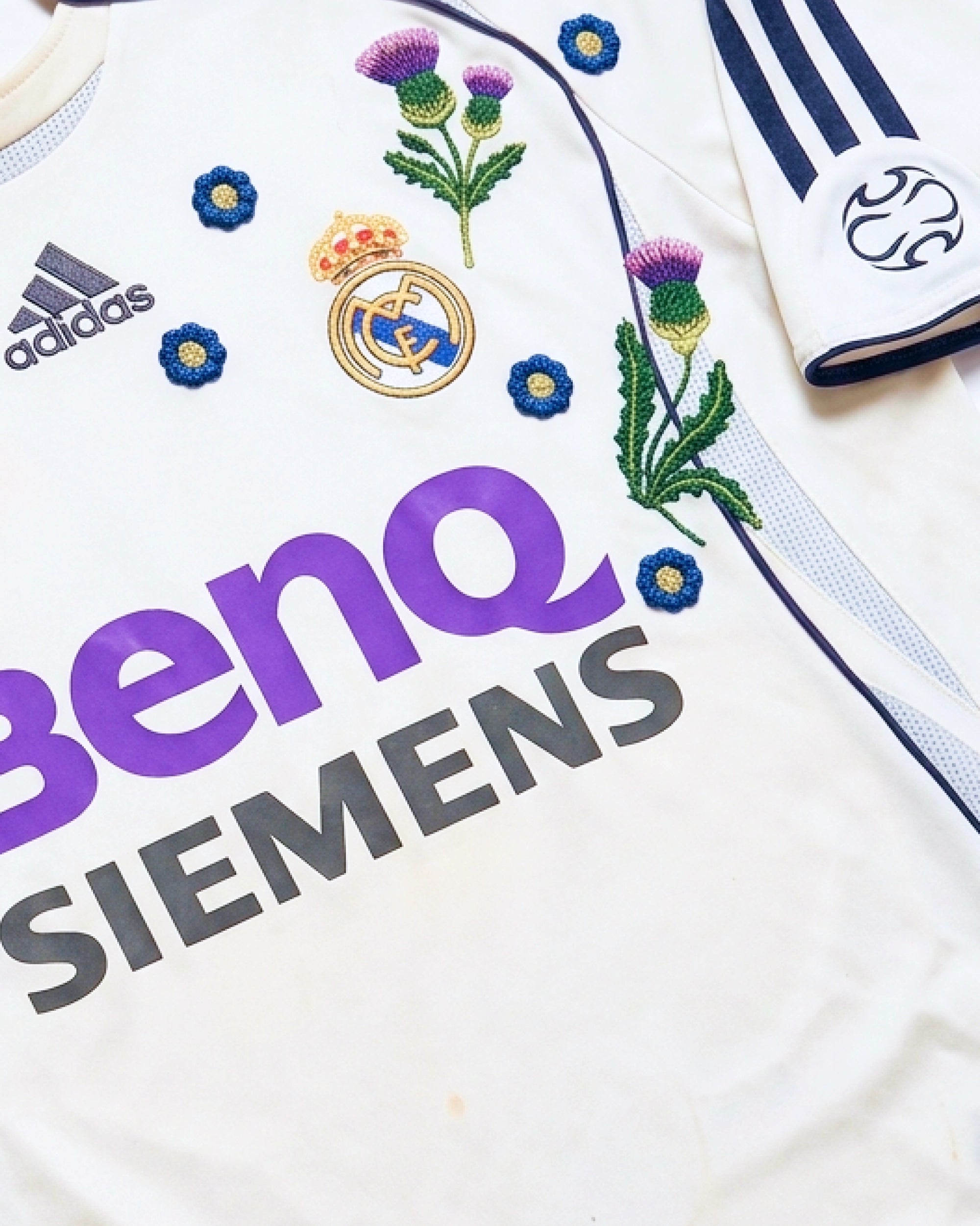 Close-up hand-embroidered purple Scottish thistle and blue botanical flowers around Real Madrid crest on retro concept jersey — artisanal embroidery VOLTA Paris & Versailles