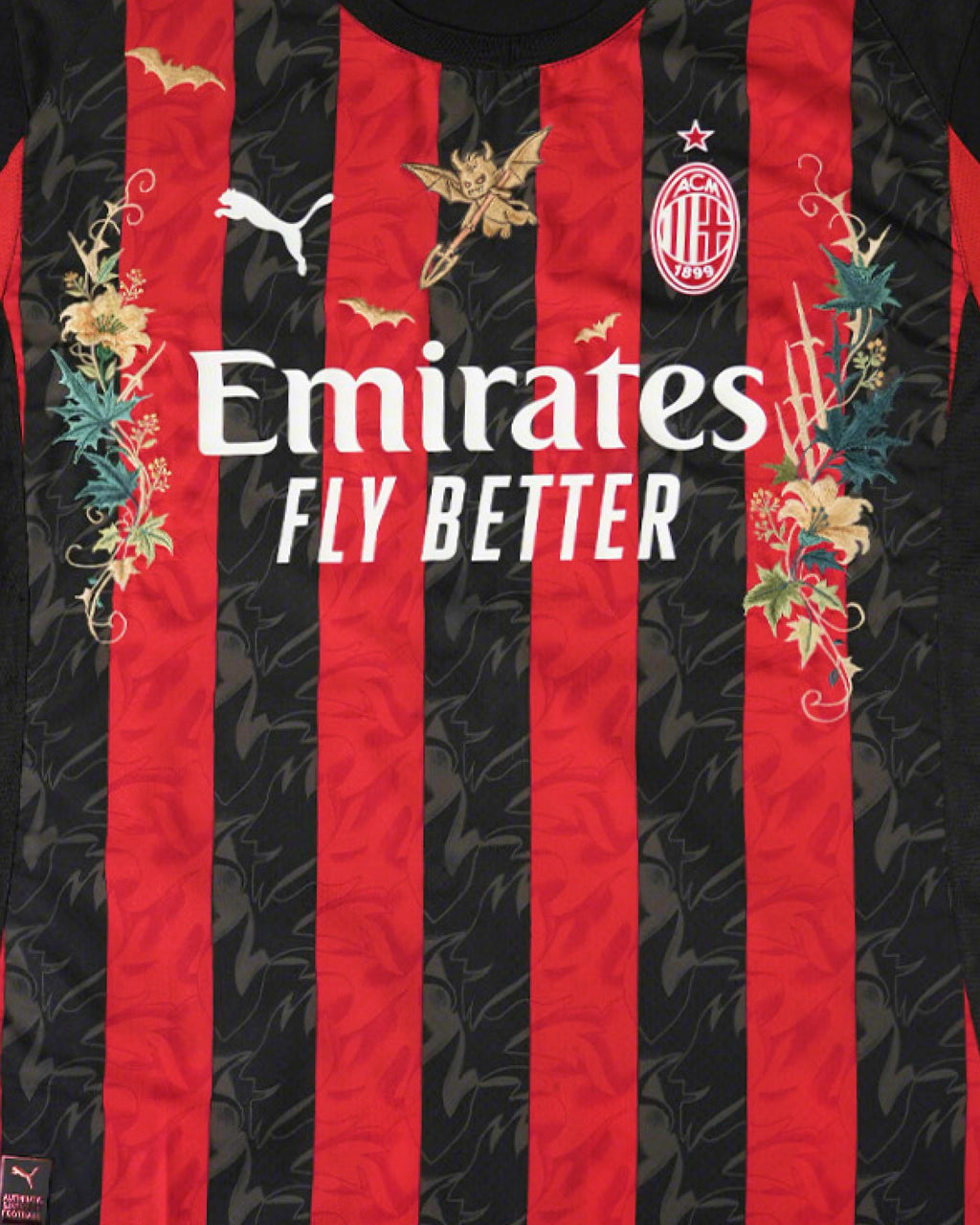 Medium Chest Shot Detail Of The AC Milan 2024/25 Jersey Reimagined, Showcasing Golden Devil, Bat, And Green Thistle Embroidery Winding Symmetrically Around The Sponsor.