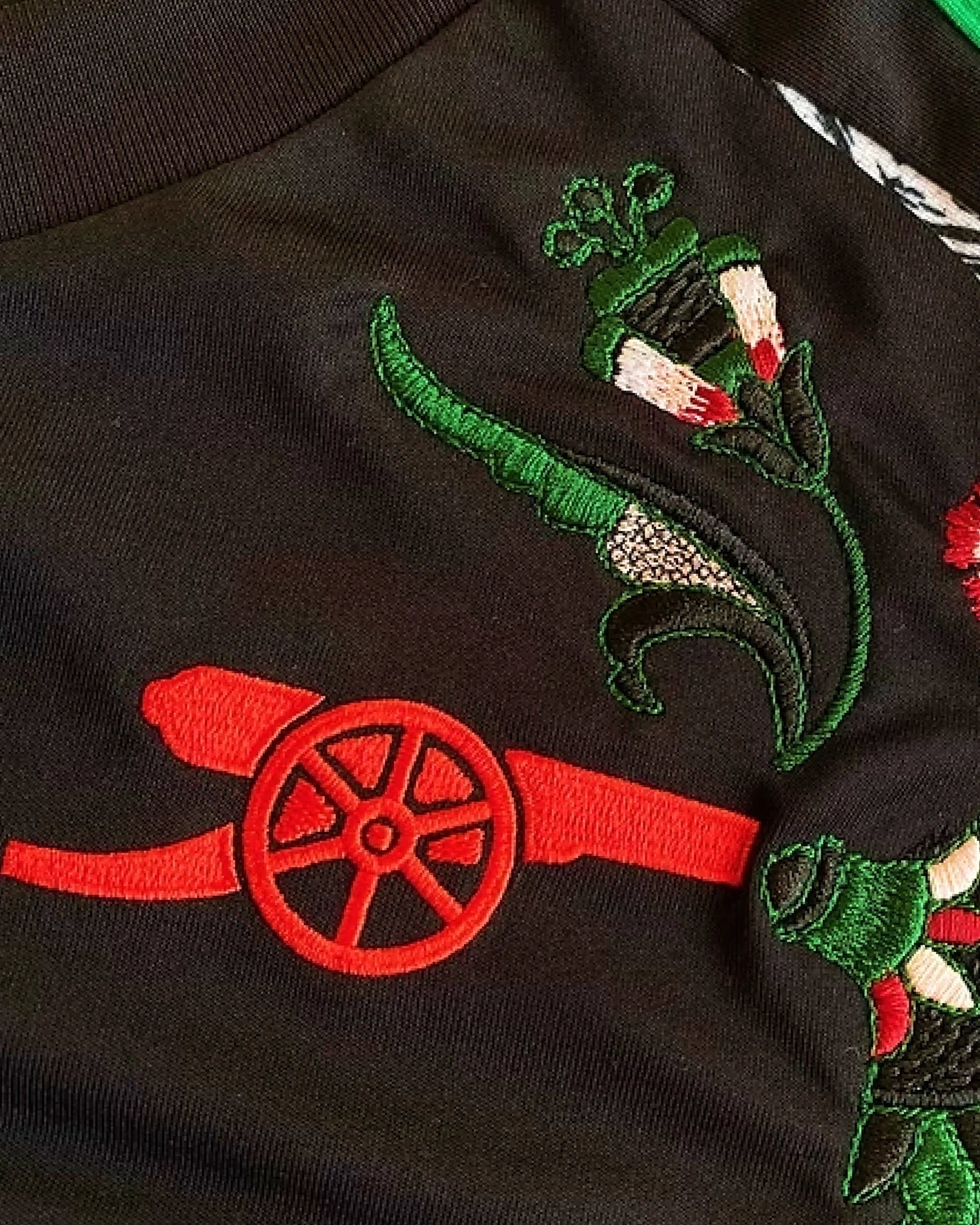 Close-Up Macro Shot Of the Left Chest On the Arsenal 2024/25 Jersey Reimagined, Highlighting The Premium Embroidered Red Cannon Crest Below Textured Green Botanical Vines.