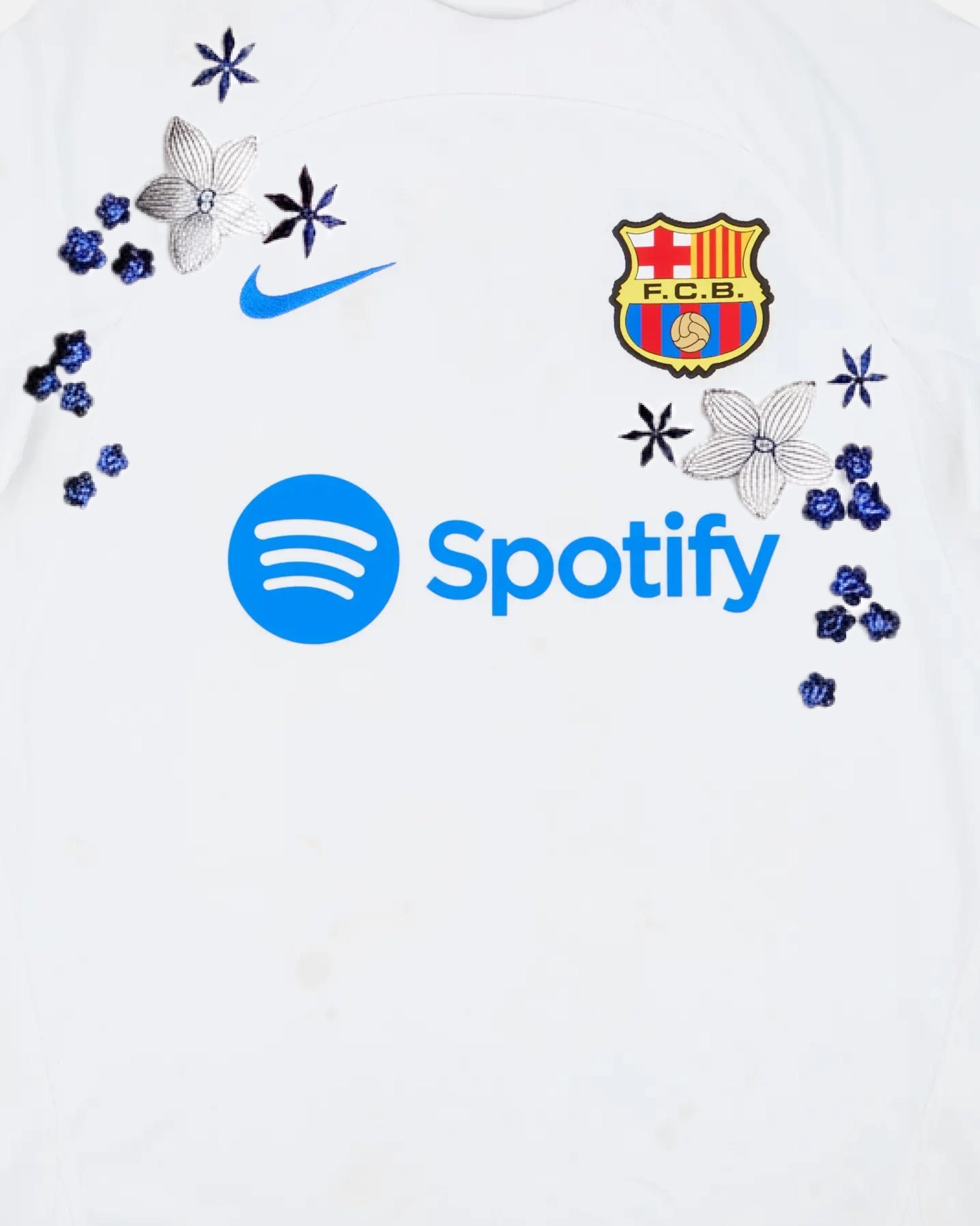 Medium Chest Shot Detail Of The Barcelona White Jersey Reimagined, Showcasing Symmetrical Silver Lily Embroidery Winding Cleanly Around The Blue Sponsor Typography.