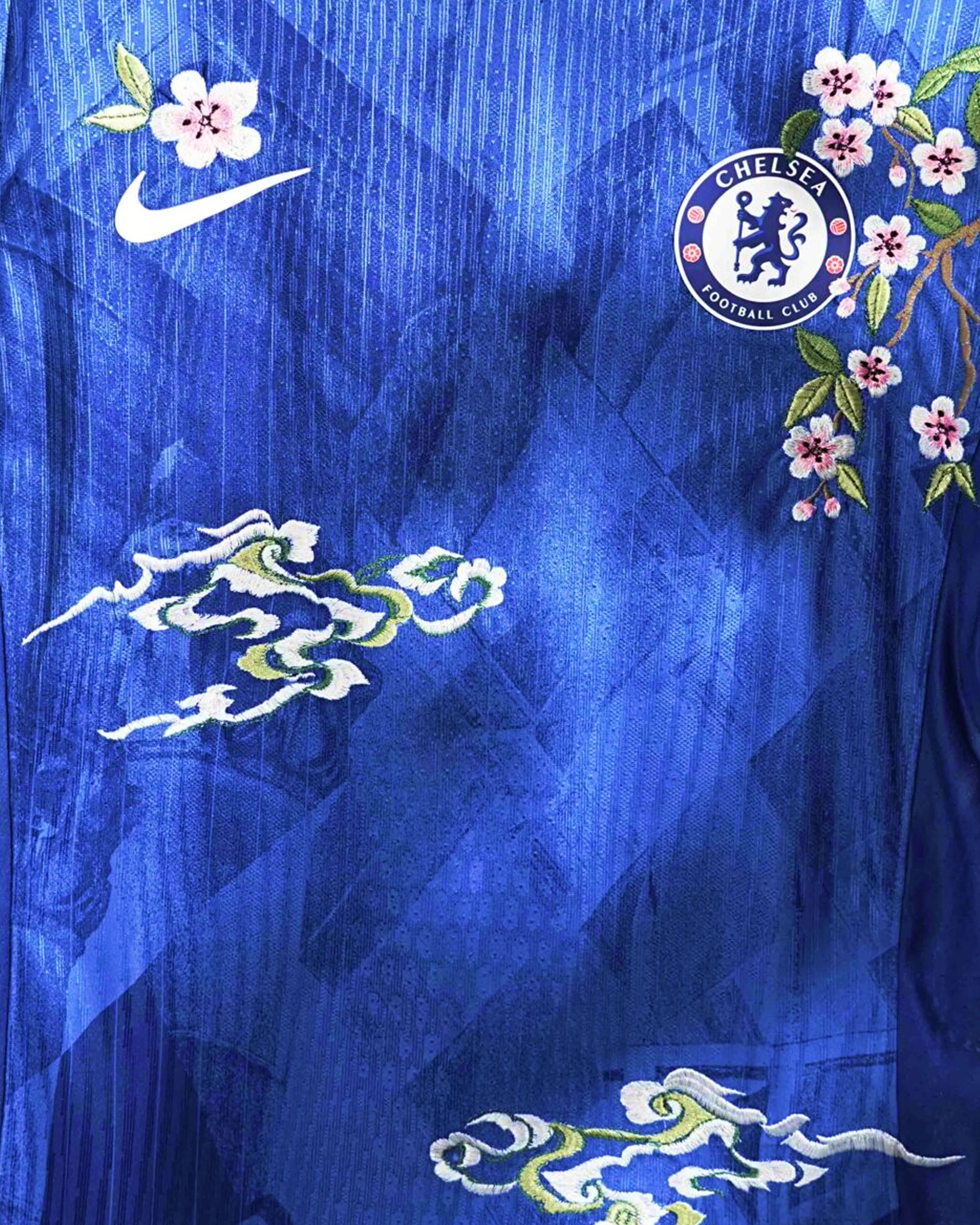 Medium Chest Shot Detail Of The Chelsea 2024/25 Jersey Reimagined, Showcasing Pink Cherry Blossom Branches Winding Around The White Crest And White Sponsor Logo.