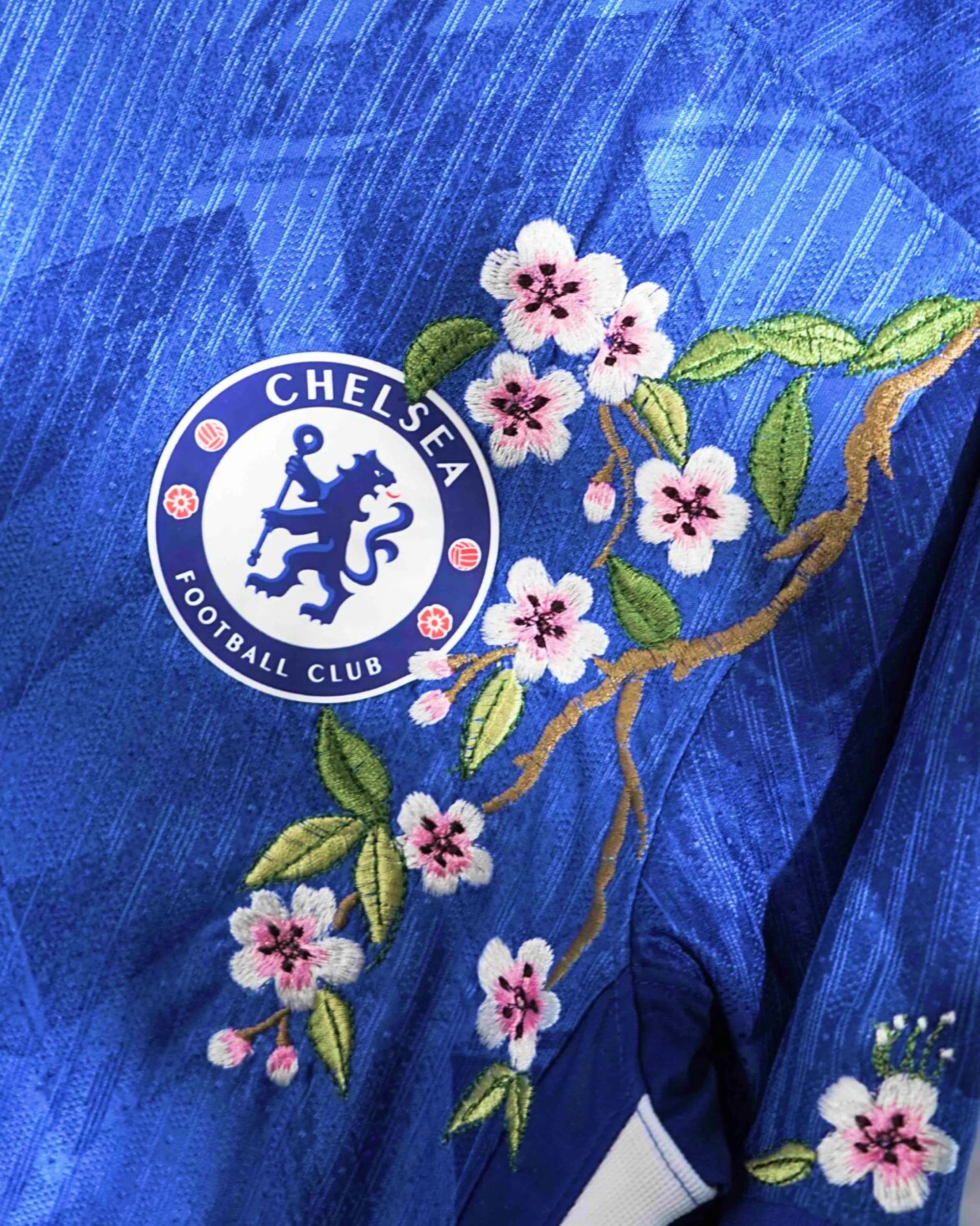 Close-Up Macro Detail On The Left Chest Of The Chelsea Home Kit Revisited, Highlighting The Dense Haptic 3D Thread Quality Of Pink Cherry Blossoms Framing The Circular Team Emblem.