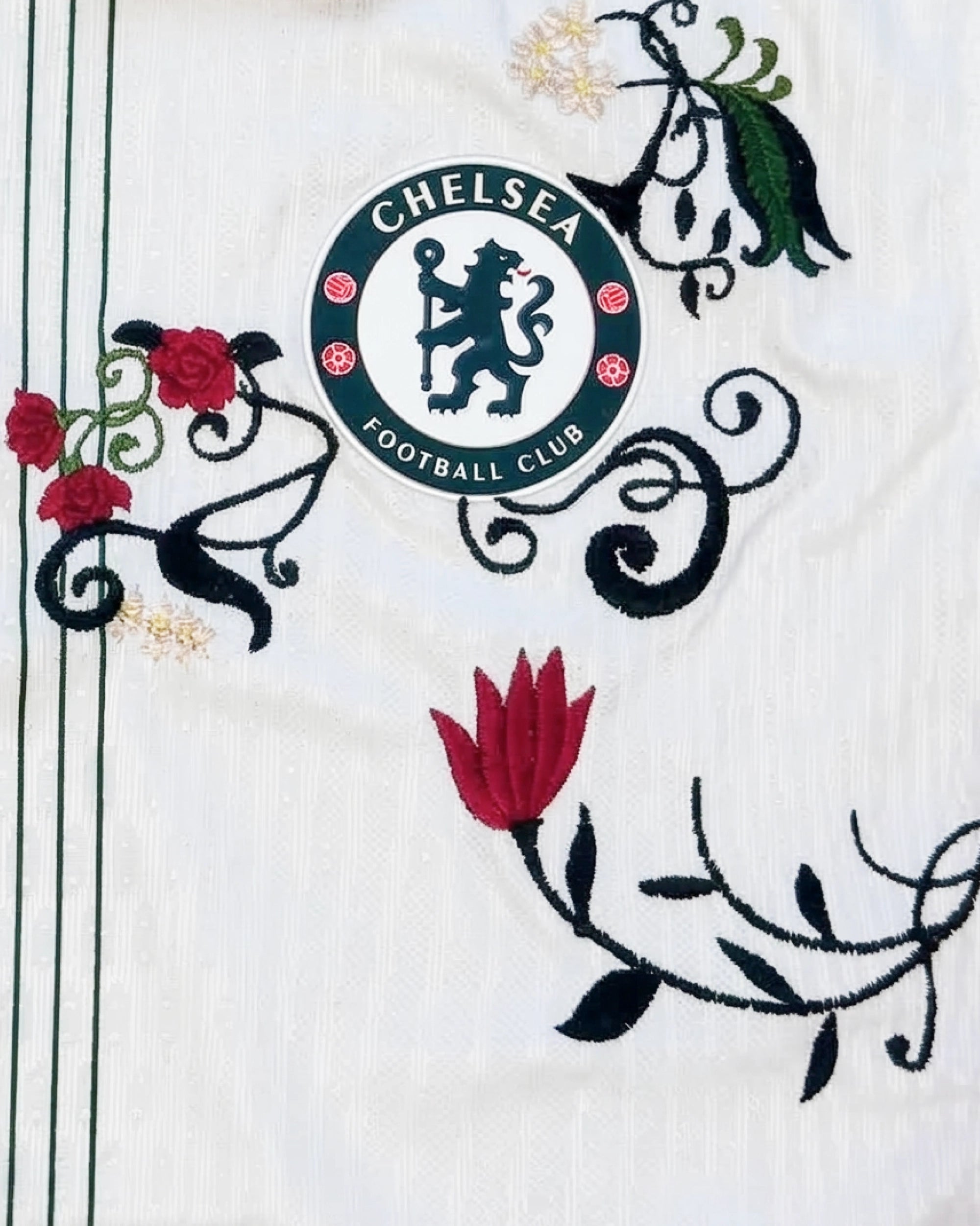 Medium Chest Shot Detail Of The Chelsea White Jersey Reimagined, Showcasing Crimson Tulip and Black Baroque Embroidery Framing The Circular Forest Green Team Crest.