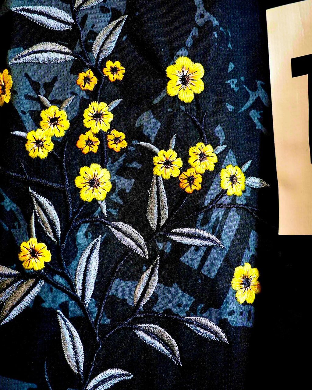 Macro Texture Shot Focusing On the Right Lower Torso Of the Custom Embroidered Black Dortmund Shirt, Highlighting The Dense Haptic 3D Thread Quality Of Yellow Wildflowers and Grey Stems.