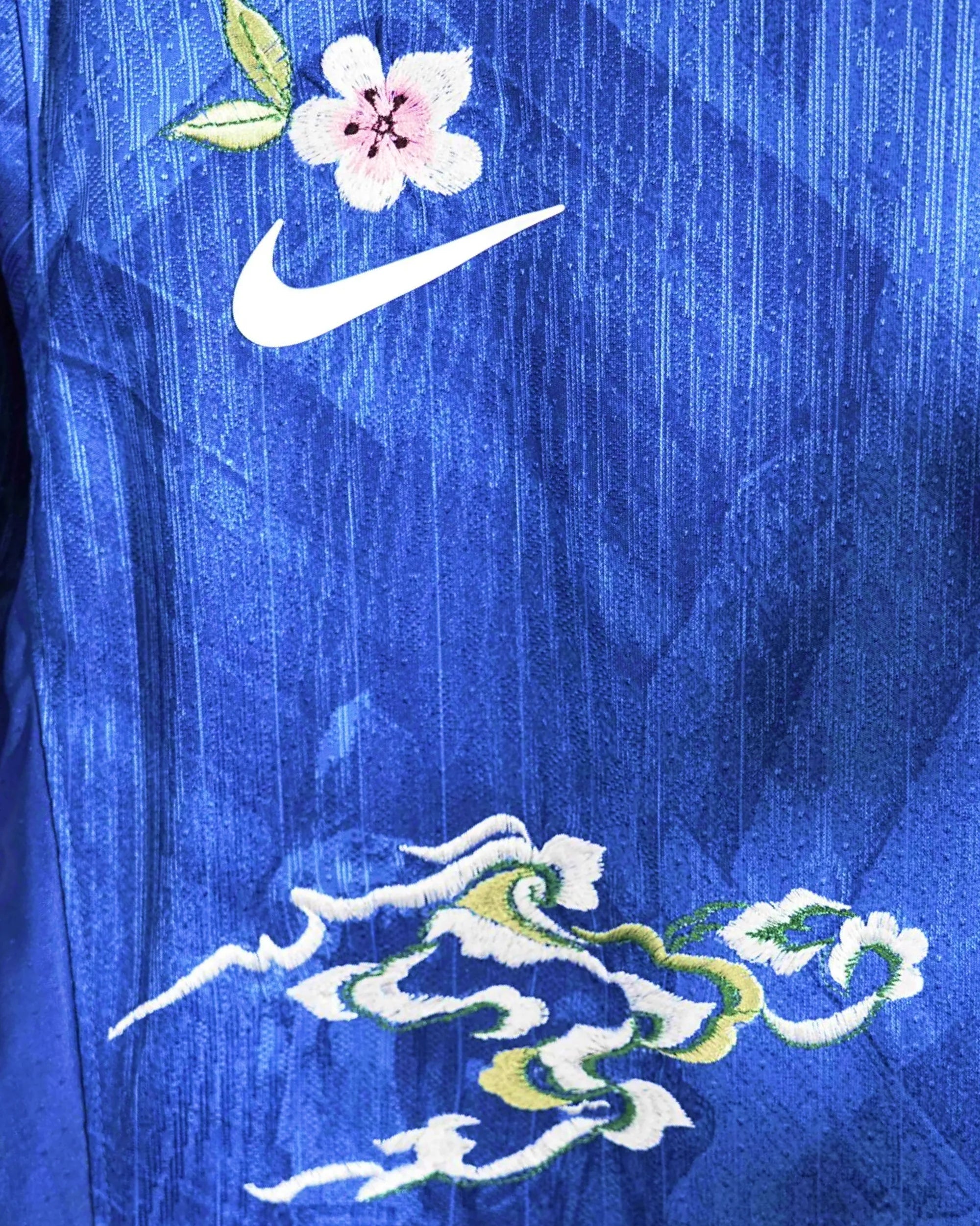 Macro Texture Shot On The Right Chest Of The Custom Embroidered Blue Chelsea Shirt, Showcasing Elegant Hand-Guided White Blossoms and Sea Clouds Next To The White Brand Logo.