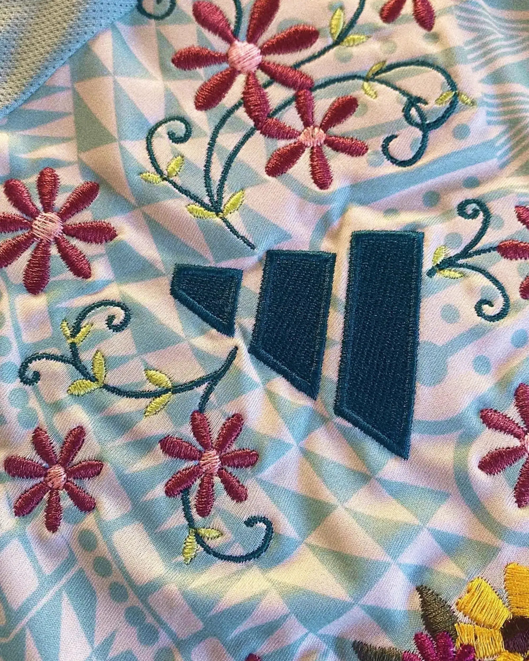 Macro Texture Shot Of the Left Chest On The Custom Embroidered Teal Football Shirt, Showcasing Elegant Hand-Guided Magenta Blossoms Enfolding The Dark Green Brand Logo.