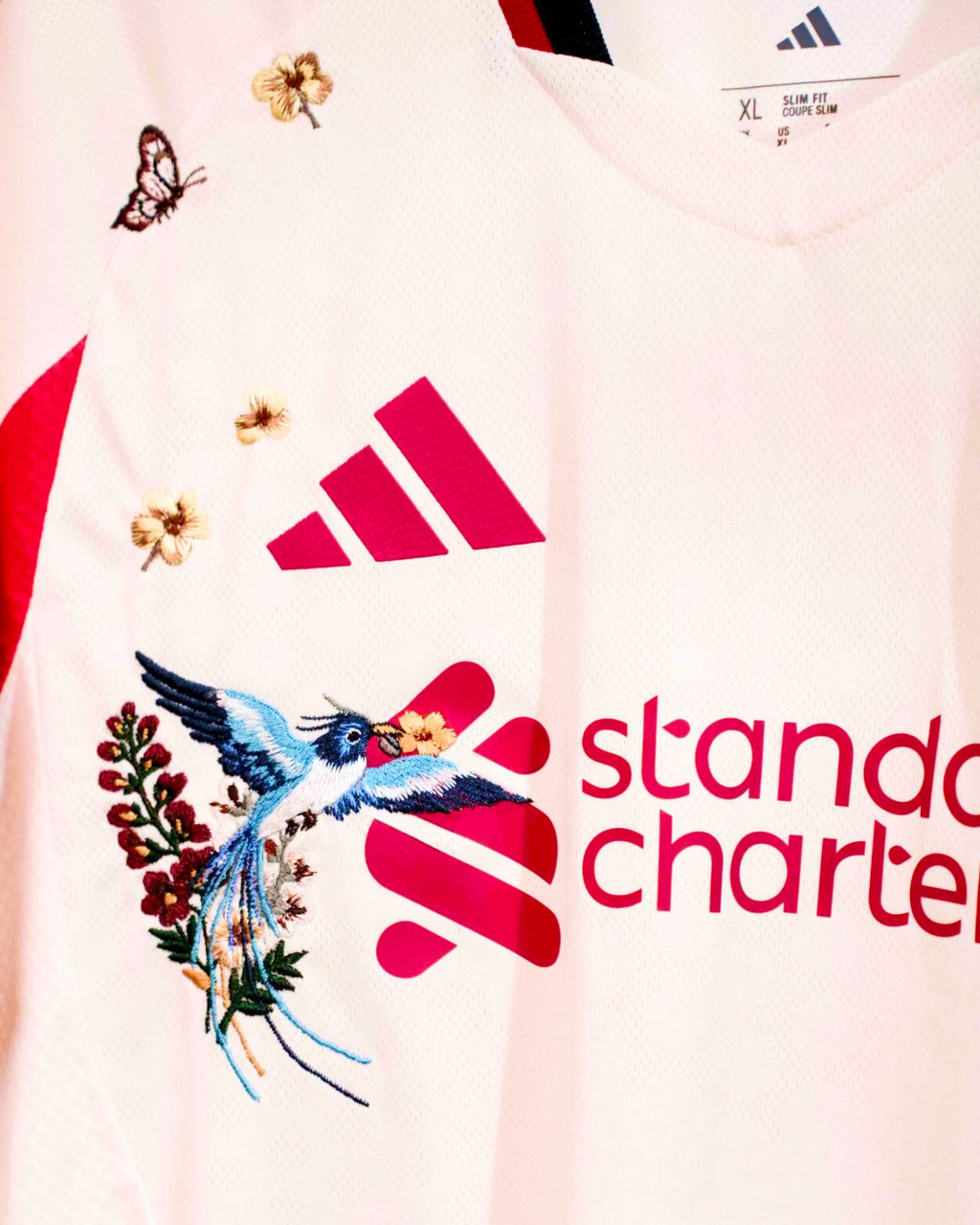 Macro Texture Shot Of The Right Chest On The Custom Embroidered White Football Shirt, Highlighting The Haptic 3D Thread Quality Of A Flying Blue Kingfisher Bird and Golden Blossoms.