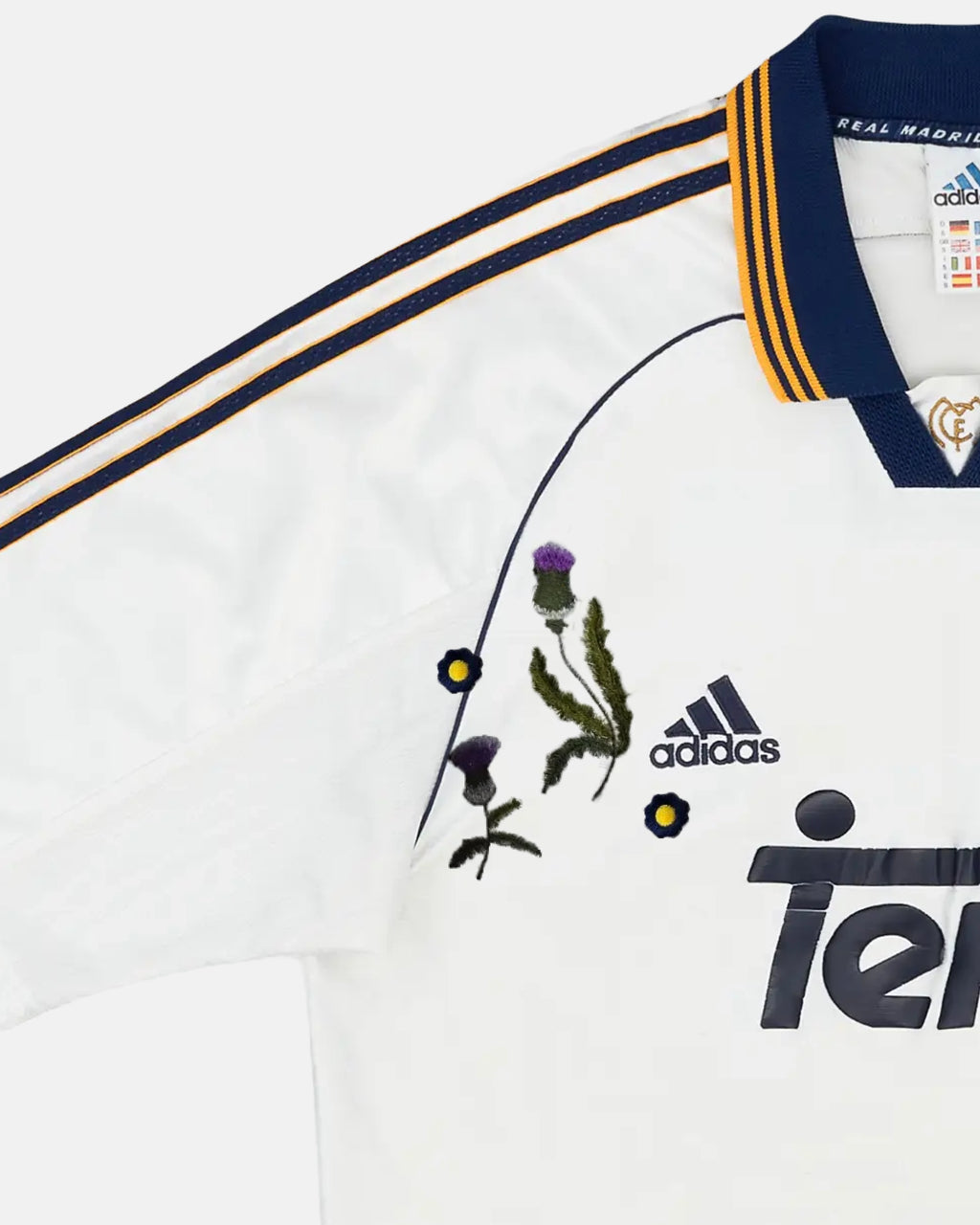 Macro Texture Shot Of The Left Chest On The Custom Embroidered White Real Madrid Shirt, Highlighting High-Density Purple Thistles Stitched Over The Dark Retro Brand Logo.