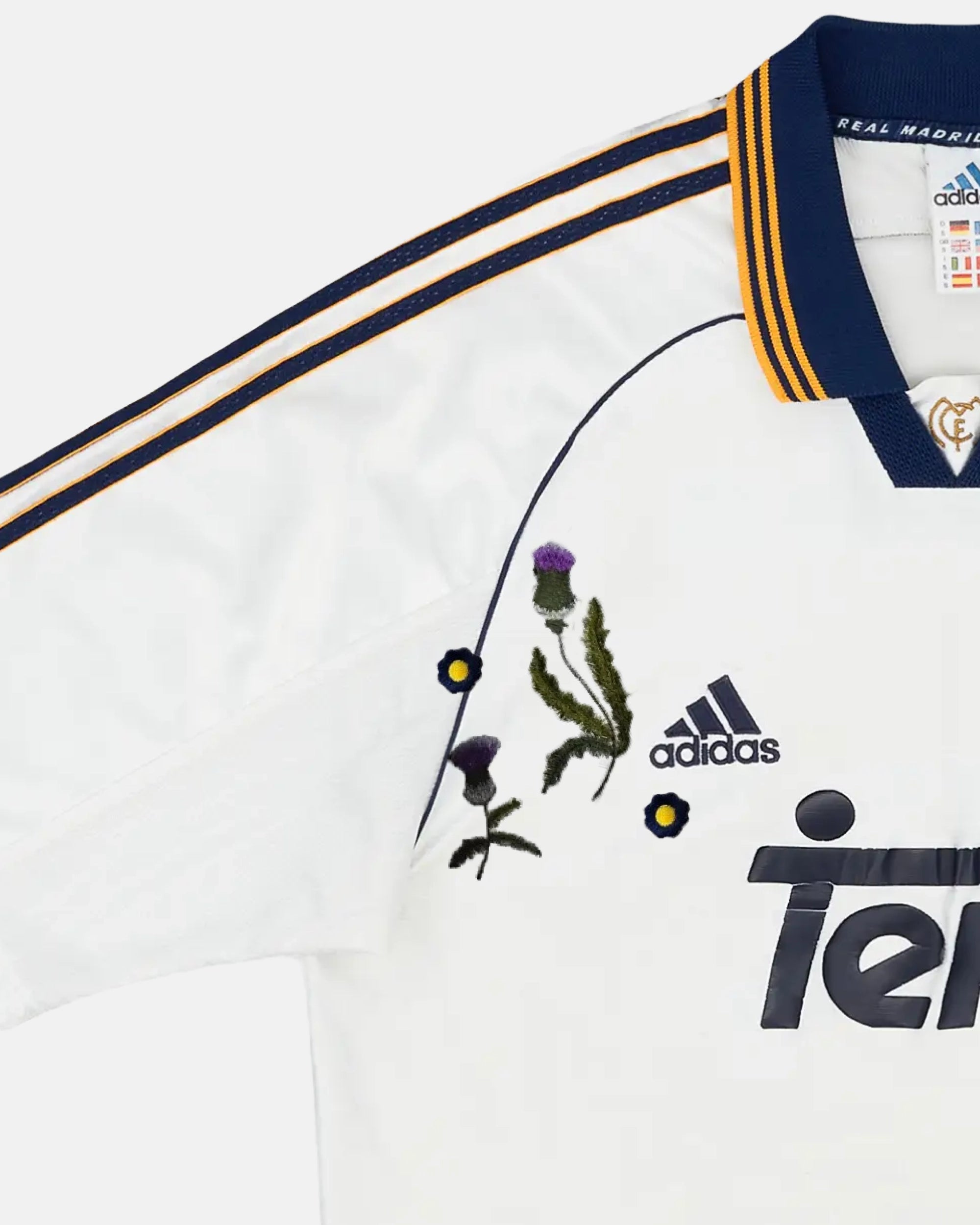 Macro Texture Shot Of The Left Chest On The Custom Embroidered White Real Madrid Shirt, Highlighting High-Density Purple Thistles Stitched Over The Dark Retro Brand Logo.