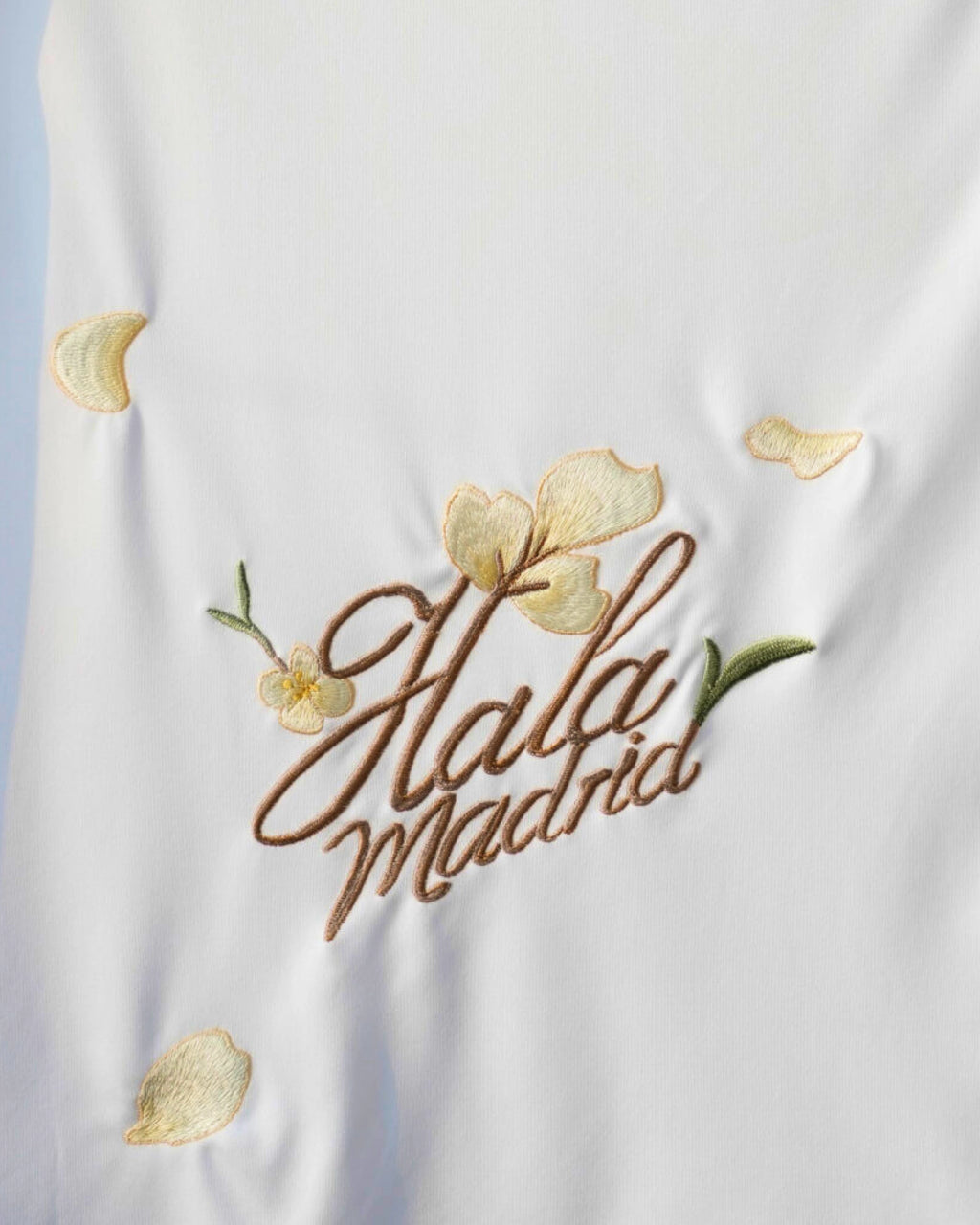 Macro Texture Shot Focusing On the Lower Rear Of the Custom Embroidered White Real Madrid Shirt, Showcasing Premium Cursive Hala Madrid Gold Stitching and Loose Petals.