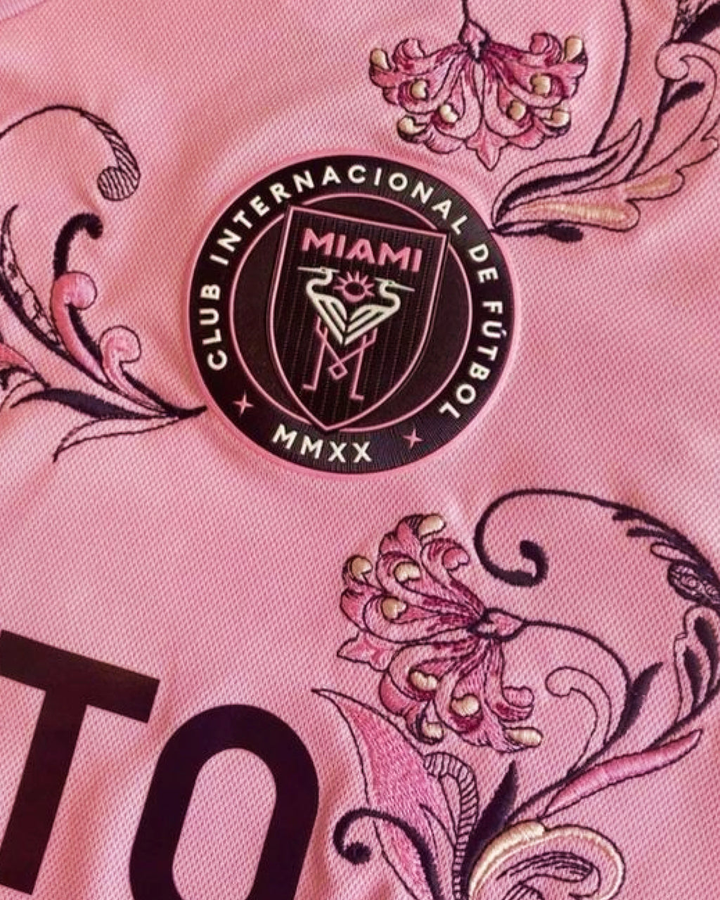 Close-Up Detail Of The Inter Miami Embroidered Pink Jersey Crest, Featuring Intricate Plum and Mauve Botanical Embroidery Surrounding The Circular Team Emblem.