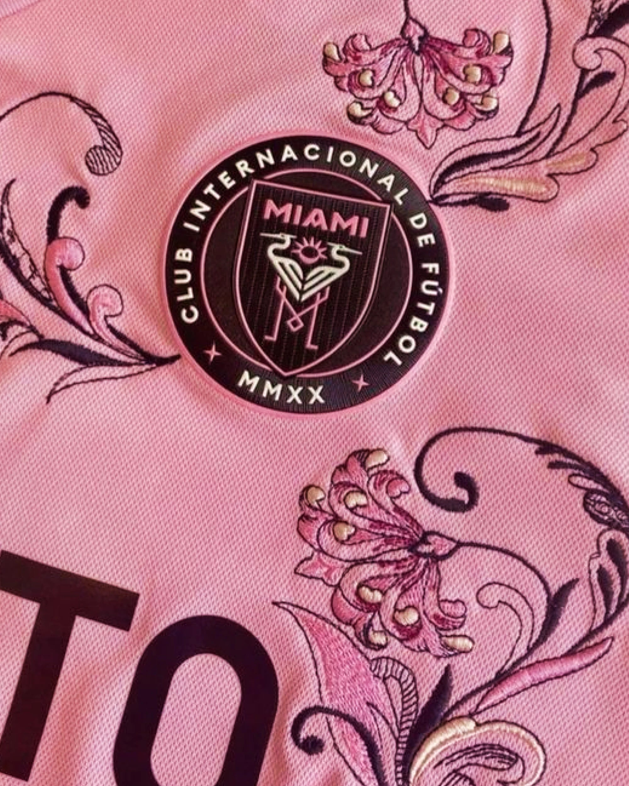 Close-Up Detail Of The Inter Miami Embroidered Pink Jersey Crest, Featuring Intricate Plum and Mauve Botanical Embroidery Surrounding The Circular Team Emblem.