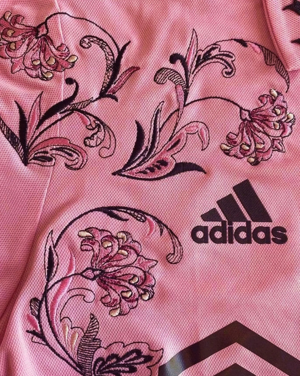 Macro Texture Shot Of The Right Chest On The Inter Miami Pink Jersey Reimagined, Showcasing The High-Density Plum Floral Embroidery Adjacent To The Black Brand Logo.