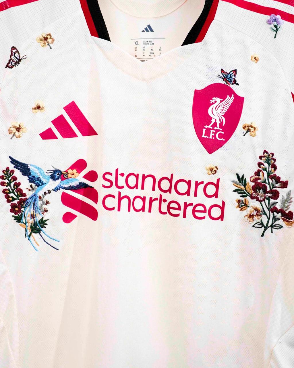 Medium Chest Shot Detail Of The Liverpool 2024/25 Jersey Reimagined, Showcasing Pristine Hand-Guided Embroidery Of A Blue Bird And Crimson Wildflowers Flanking The Sponsor Font.