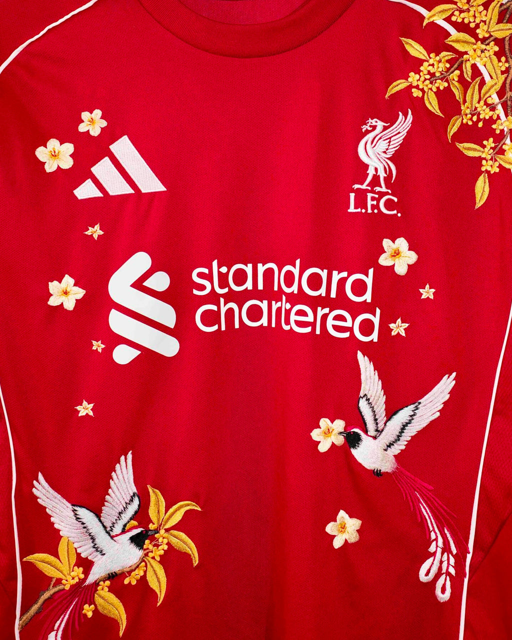 Medium Chest Shot Detail Of The Liverpool 2024/25 Jersey Reimagined, Showcasing Elegant White Birds Of Paradise And Floating Gold Flowers Flanking The Sponsor.