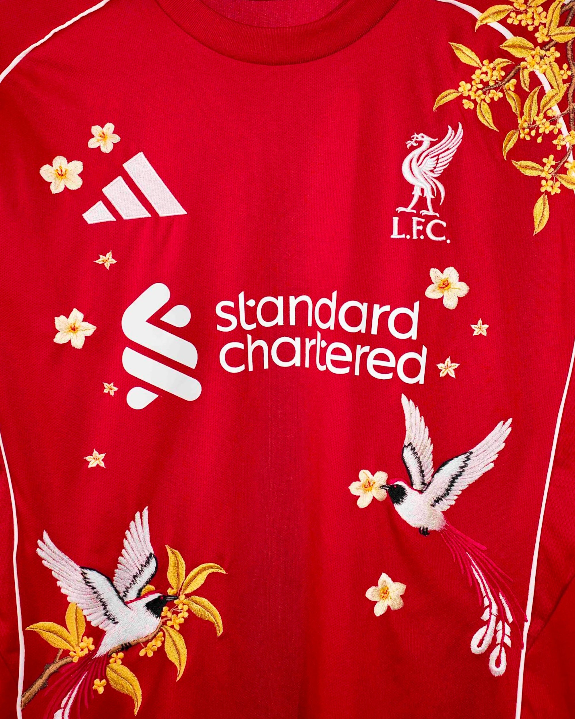 Medium Chest Shot Detail Of The Liverpool 2024/25 Jersey Reimagined, Showcasing Elegant White Birds Of Paradise And Floating Gold Flowers Flanking The Sponsor.