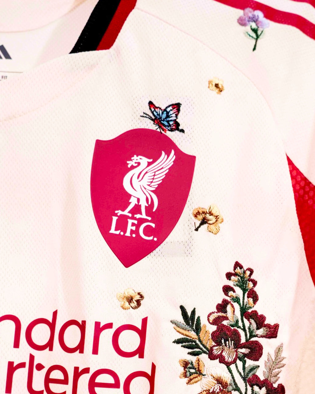 Close-Up Macro Detail Of The Liverpool Away Kit Revisited Shield Crest, Highlighting Crimson Orchid Floral Stitches and A Miniature Butterfly Balancing Next To The Liver Bird Emblem.