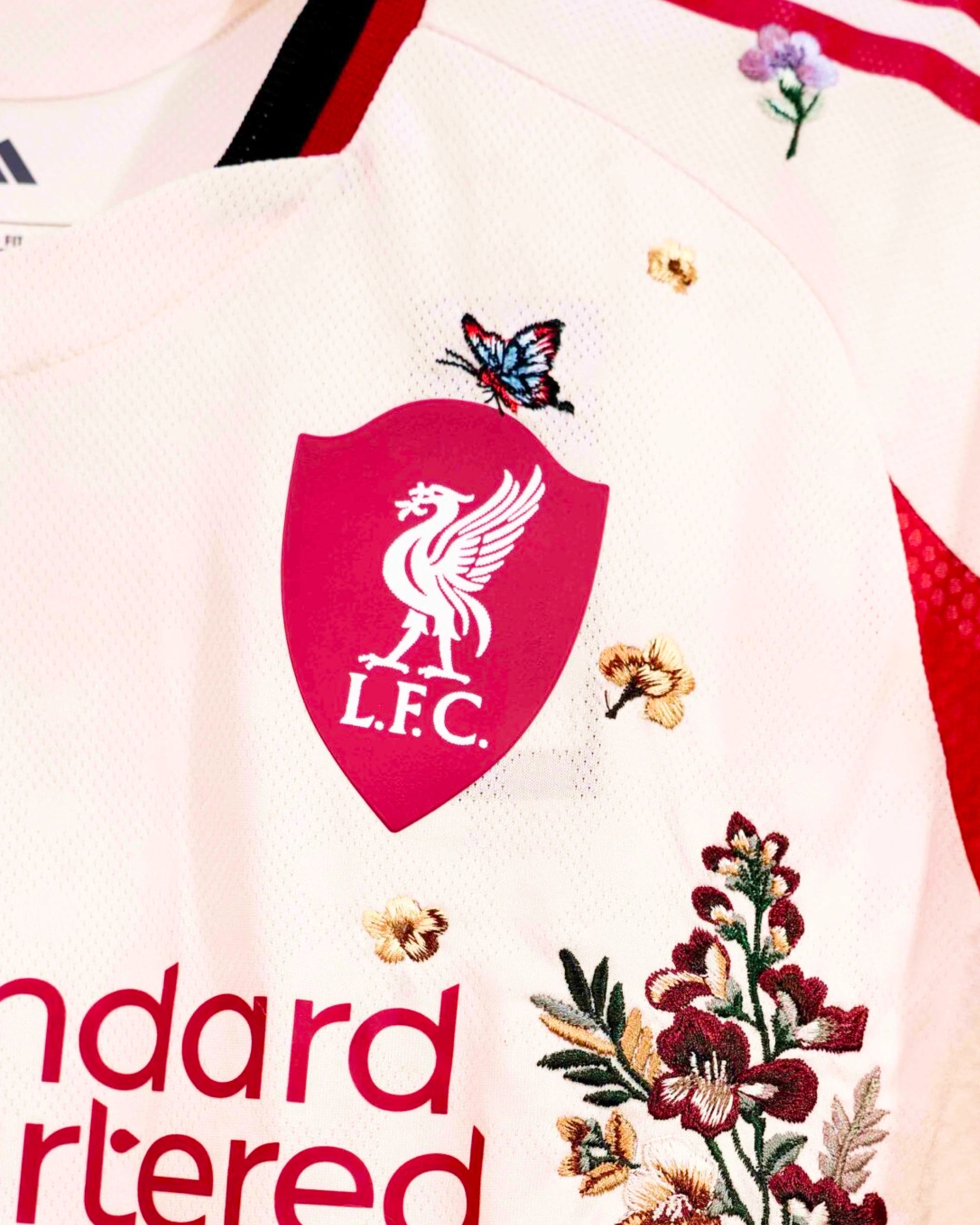 Close-Up Macro Detail Of The Liverpool Away Kit Revisited Shield Crest, Highlighting Crimson Orchid Floral Stitches and A Miniature Butterfly Balancing Next To The Liver Bird Emblem.