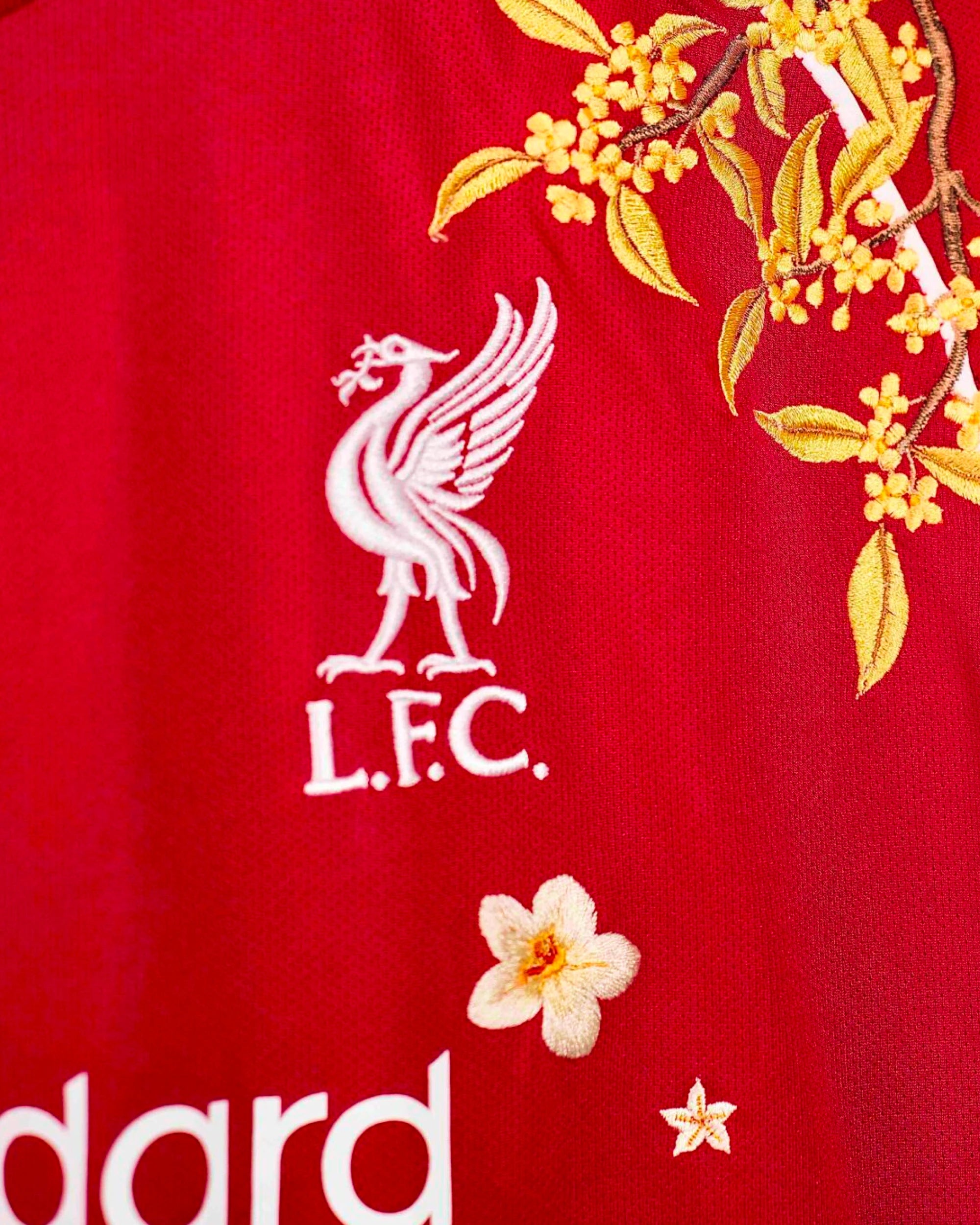 Close-Up Macro Detail On The Left Chest Of The Liverpool Home Kit Revisited, Highlighting The White Woven Liver Bird Emblem Beneath A Metallic Gold Floral Branch.