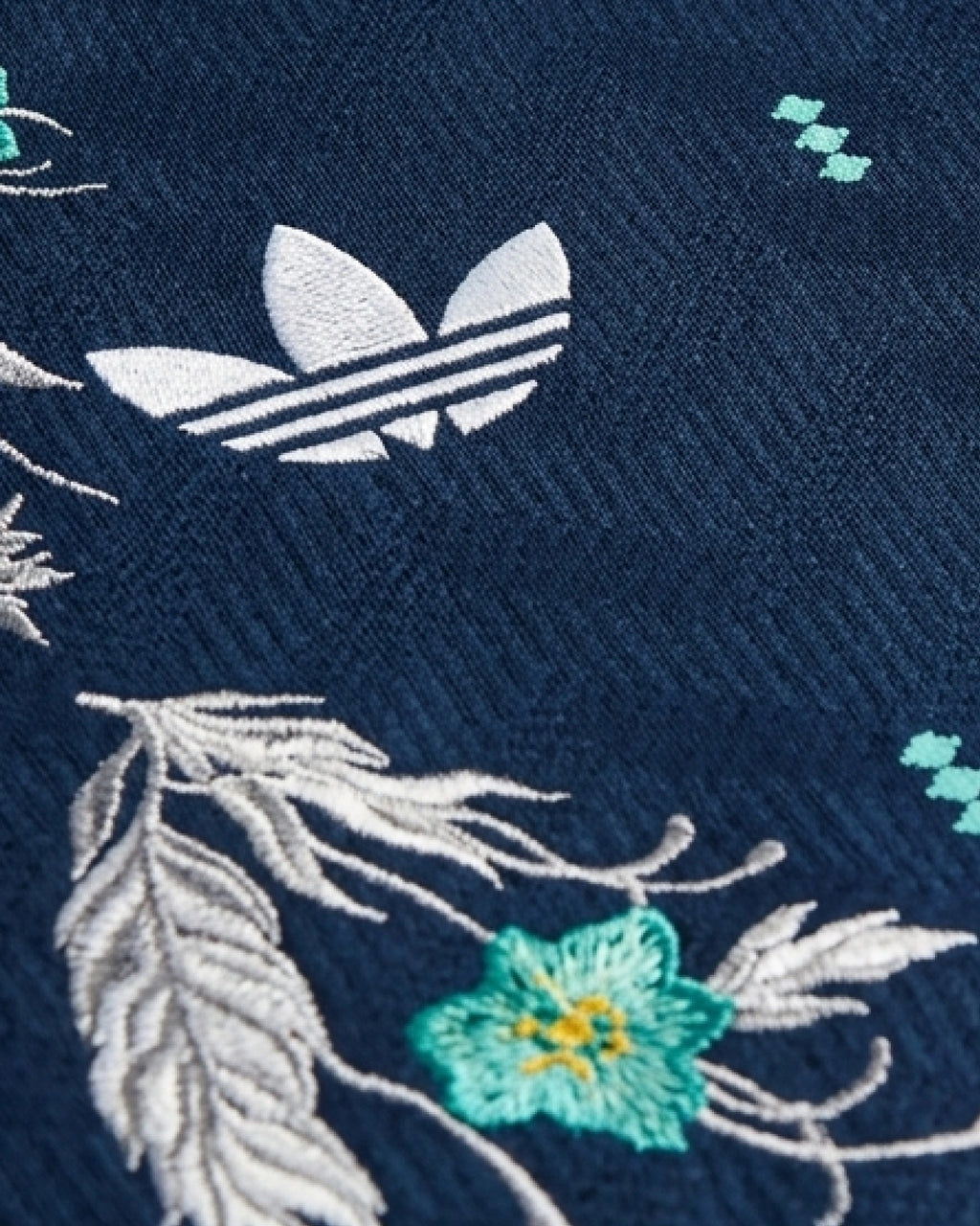 Macro Texture Zoom Lens Focus On The White Trefoil Logo, Emphasizing The Haptic 3D Embroidery Texture of Silver Leaves And Mint Petals On Navy Fabric.