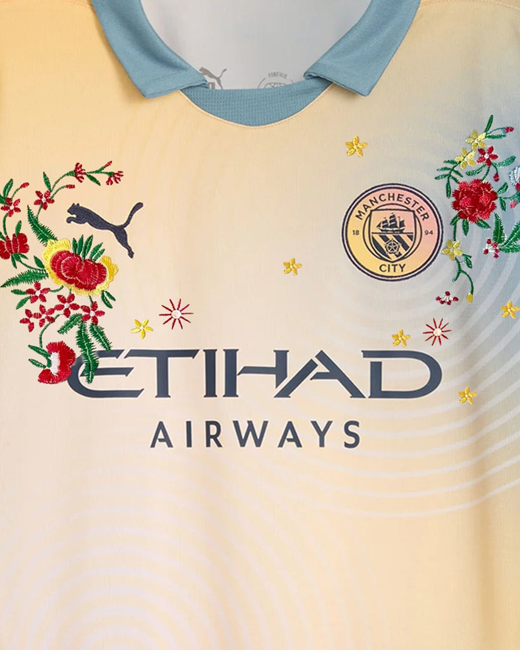 Medium Chest Shot Detail Of The Manchester City 2024/25 Jersey Reimagined, Showcasing Symmetrical Floral Embroidery Winding Seamlessly Around The Dark Navy Sponsor Text.