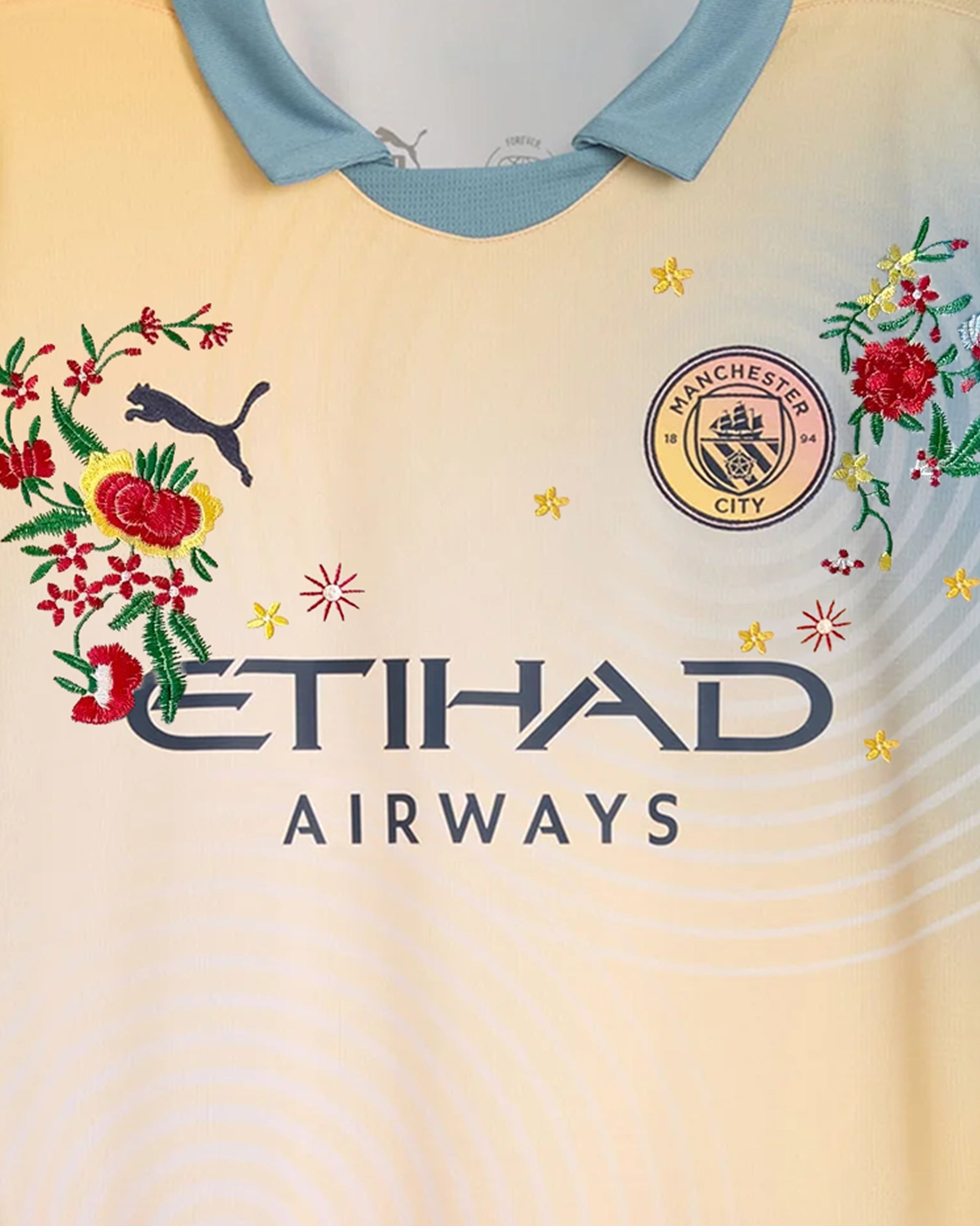 Medium Chest Shot Detail Of The Manchester City 2024/25 Jersey Reimagined, Showcasing Symmetrical Floral Embroidery Winding Seamlessly Around The Dark Navy Sponsor Text.