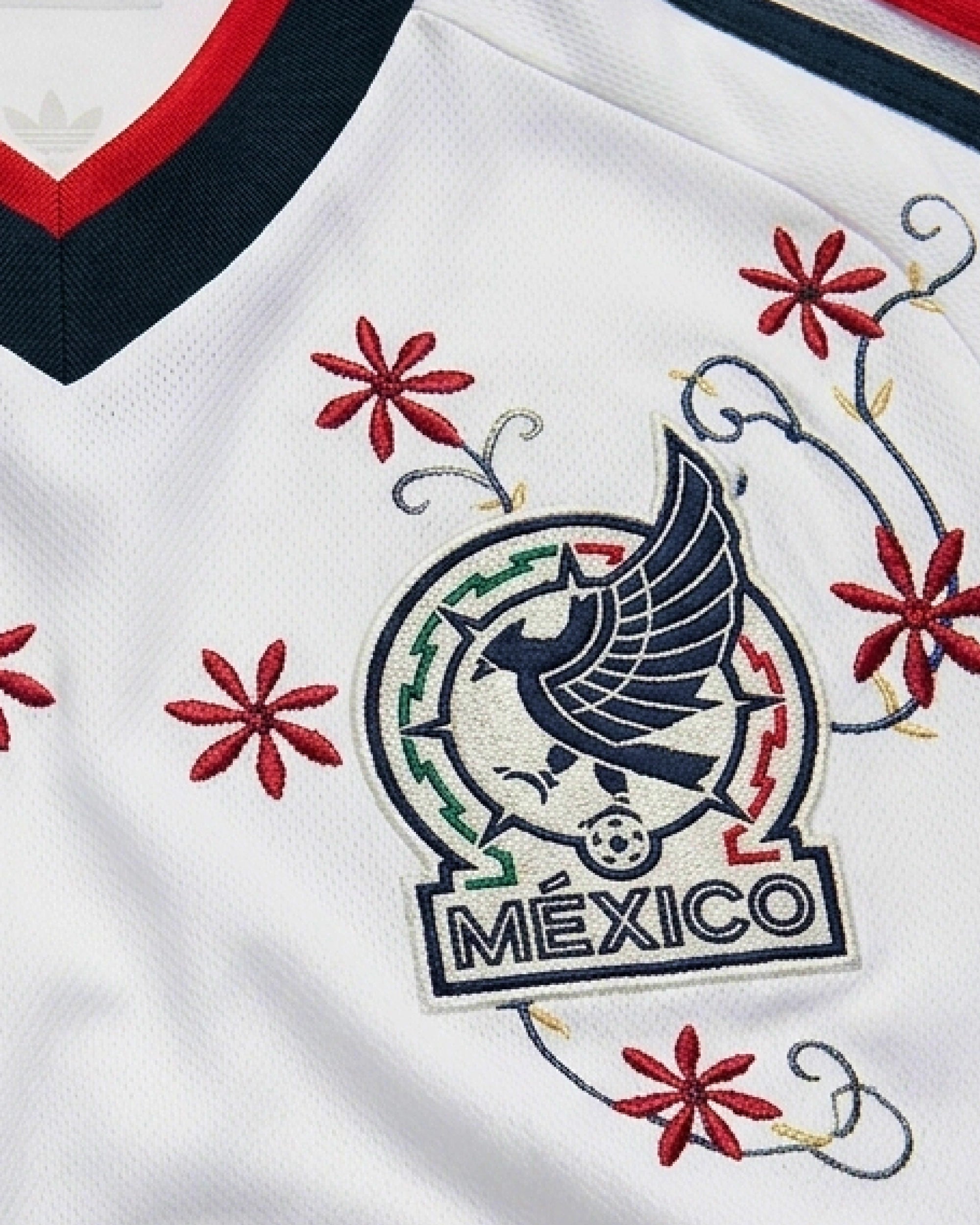 Close-Up Shot Of The Mexico World Cup Jersey Reimagined Left Chest, Displaying The Geometric Mexican Eagle Crest Enwrapped In Crimson Artisan Starburst Flowers.