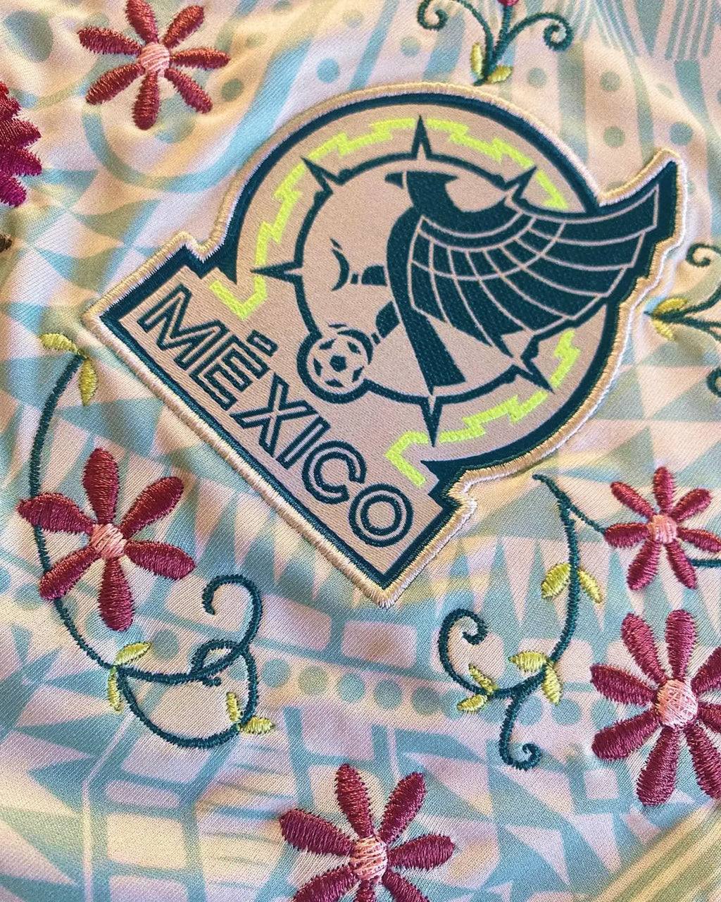 Close-Up Detail Of The Mexico World Cup Kit Revisited Team Shield, Illustrating Deep Magenta Floral Embroidery Winding Beneath The Silver And Neon Green Eagle Crest.