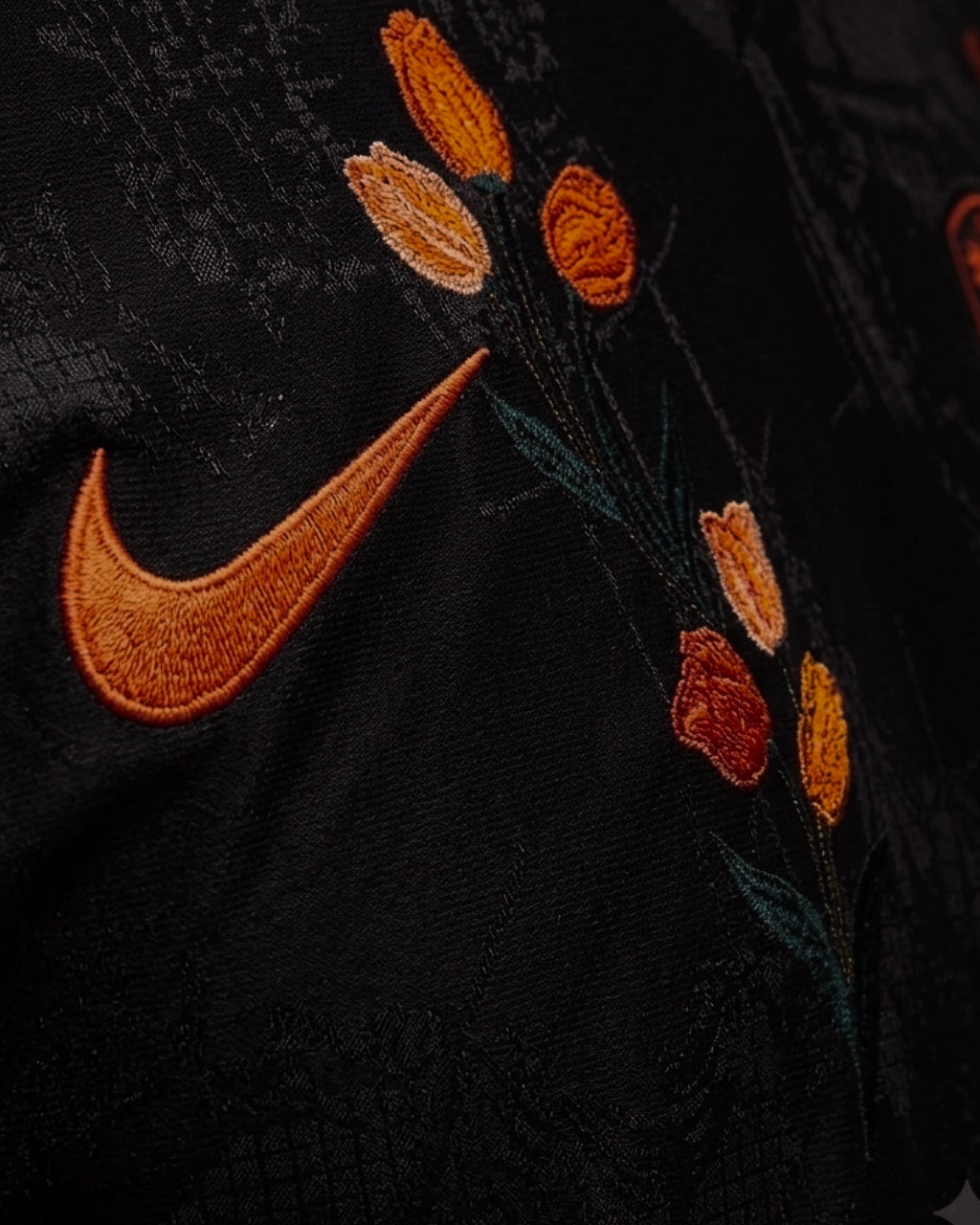 Ultra close-up hand-embroidered orange tulips in deep orange and amber thread cascading beside swoosh on Netherlands black concept jersey — artisanal botanical embroidery VOLTA Paris & Versailles