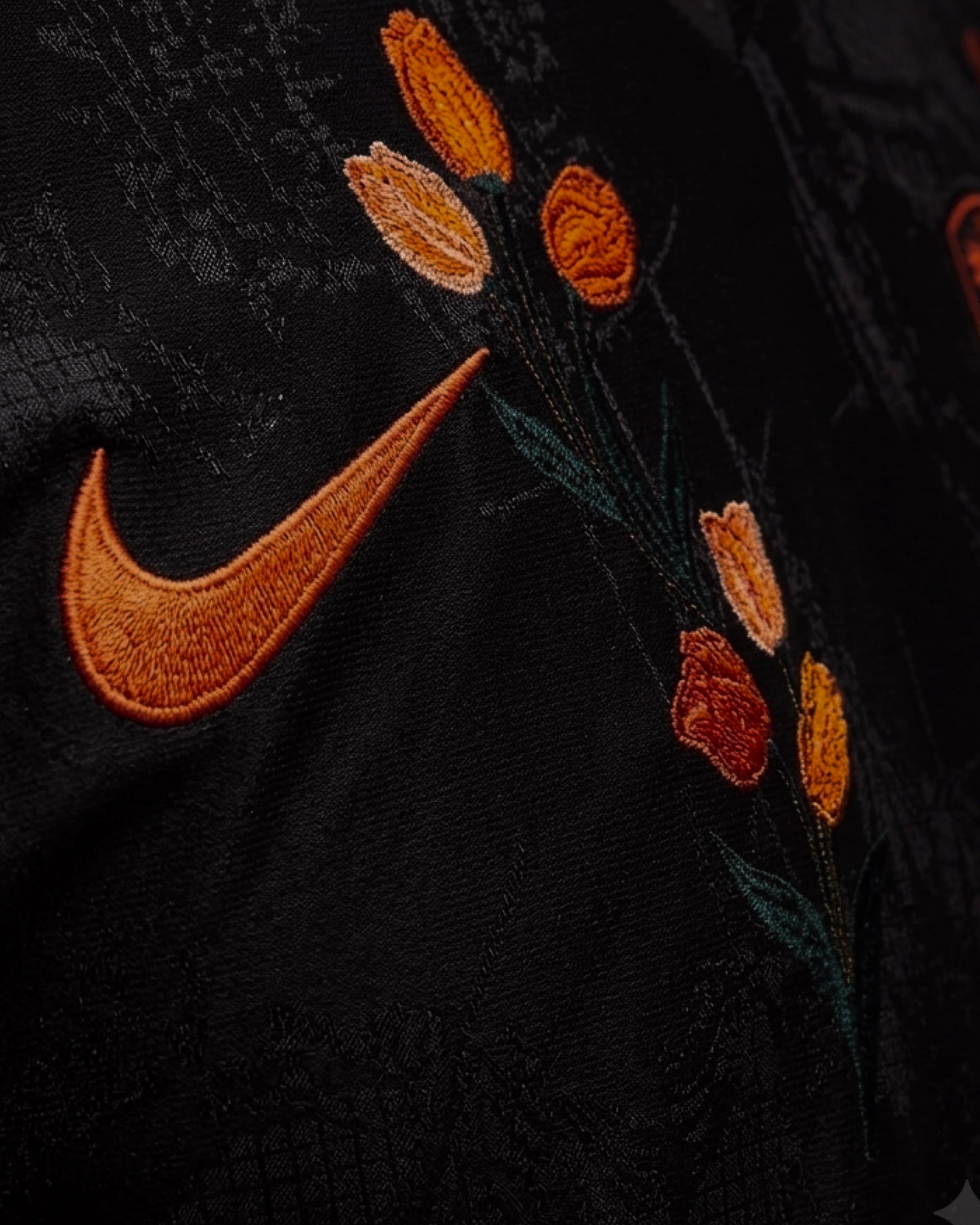 Ultra close-up hand-embroidered orange tulips in deep orange and amber thread cascading beside swoosh on Netherlands black concept jersey — artisanal botanical embroidery VOLTA Paris & Versailles