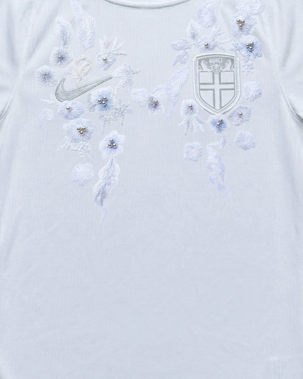 Medium shot detail of the Norway Embroidered White Jersey, illustrating how the icy-blue winter blossoms and silver-white foliage frame the national crest and silver brand logo.