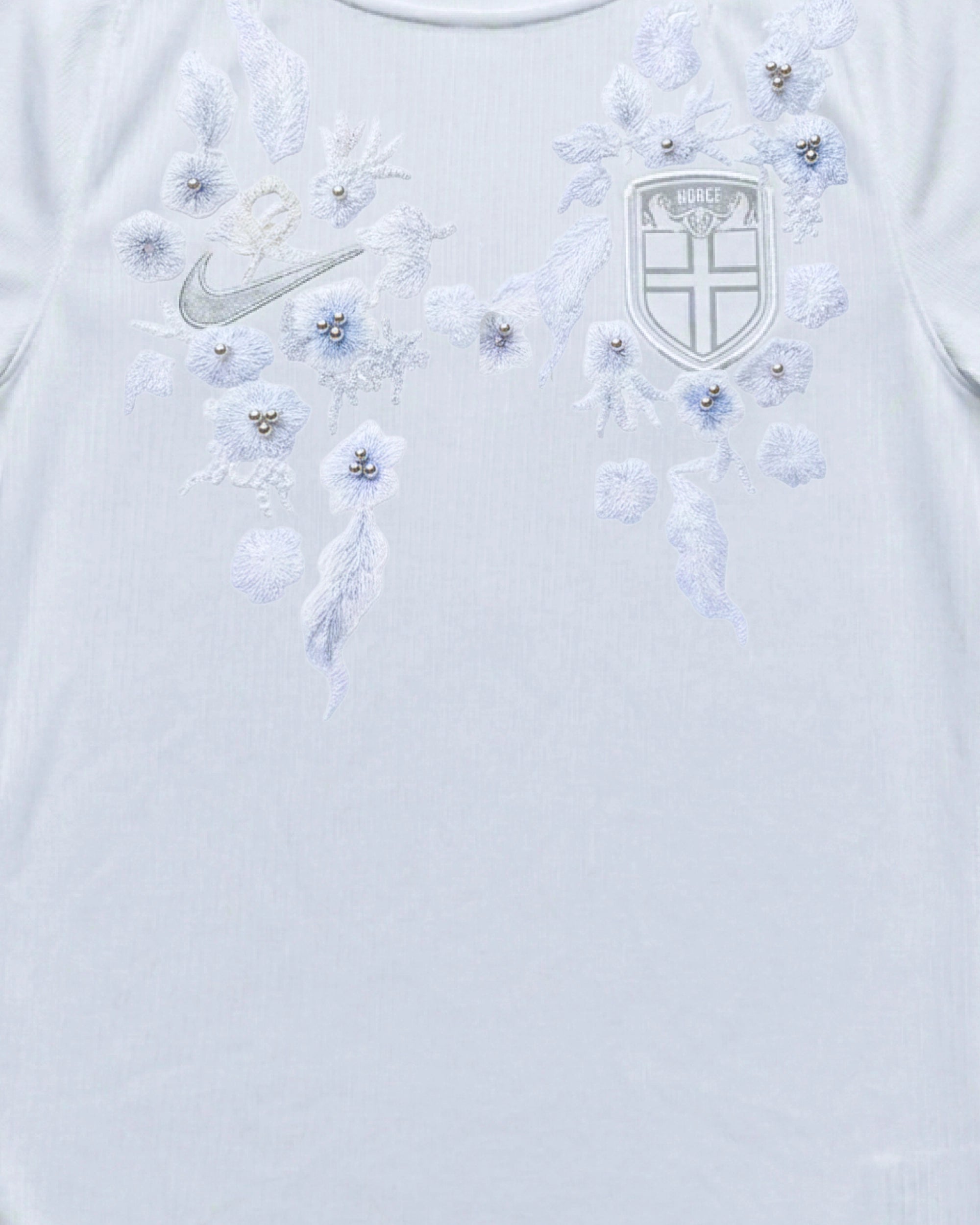 Medium shot detail of the Norway Embroidered White Jersey, illustrating how the icy-blue winter blossoms and silver-white foliage frame the national crest and silver brand logo.