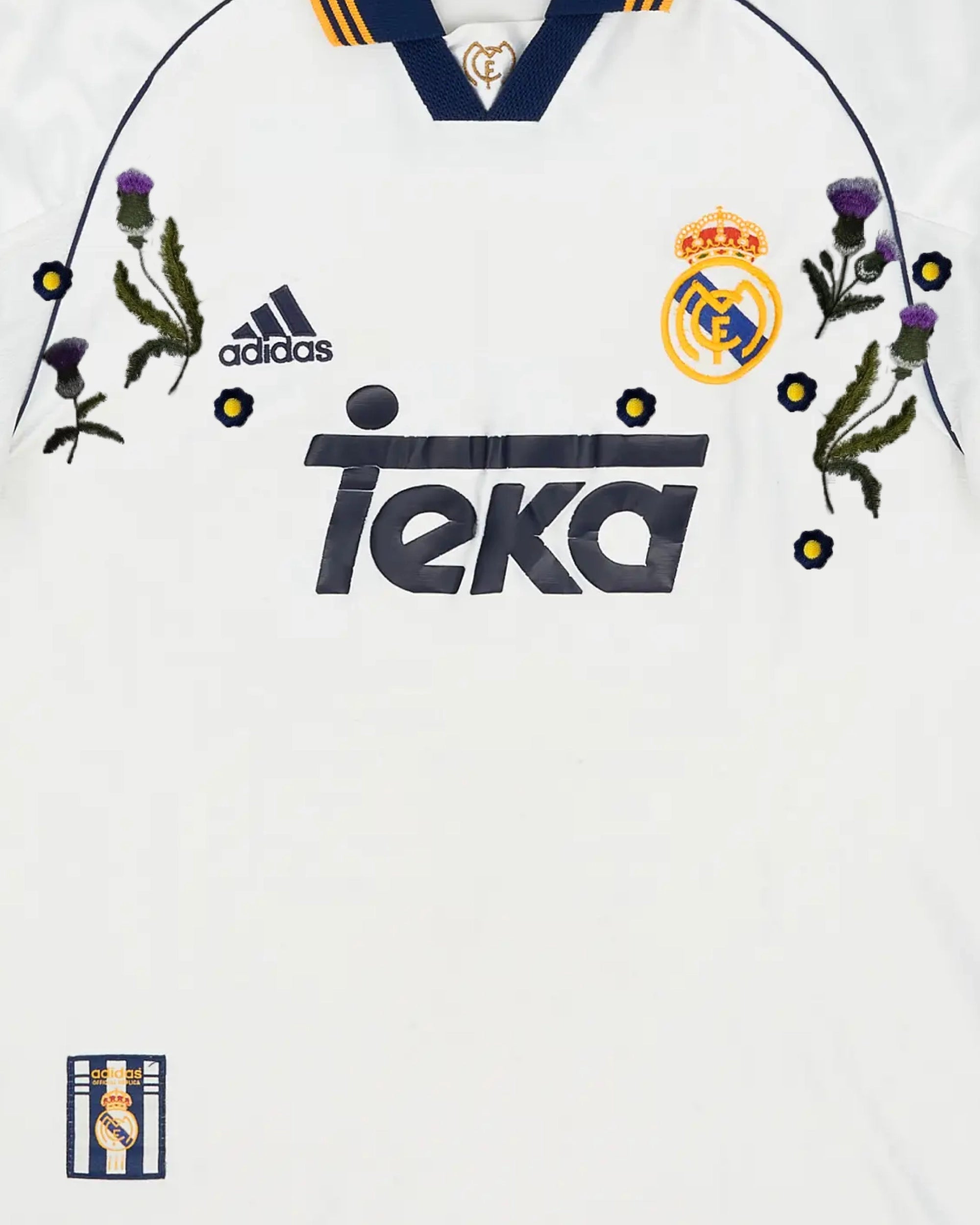 Medium Chest Shot Detail Of The Real Madrid 1999/2000 Jersey Reimagined, Showcasing Symmetrical Purple Thistle Embroidery Flanking The Raised Retro Sponsor Text.