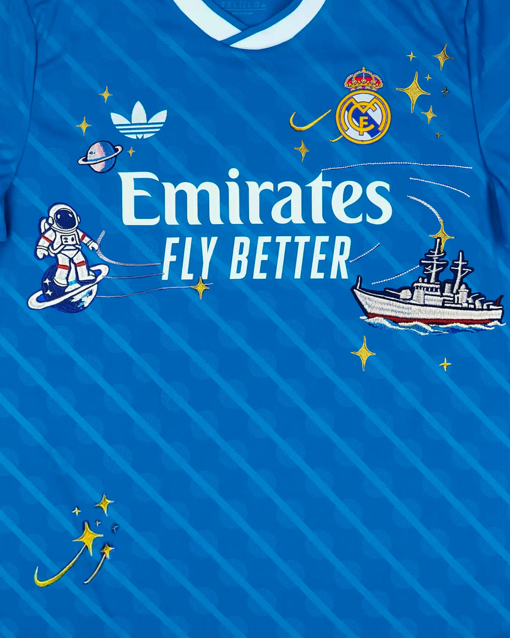 Medium Chest Shot Detail Of The Real Madrid Blue Jersey Reimagined, Showcasing A Silver Stitched Rocket Trail Connecting An Astronaut Graphic To A Naval Warship Across The Sponsor.