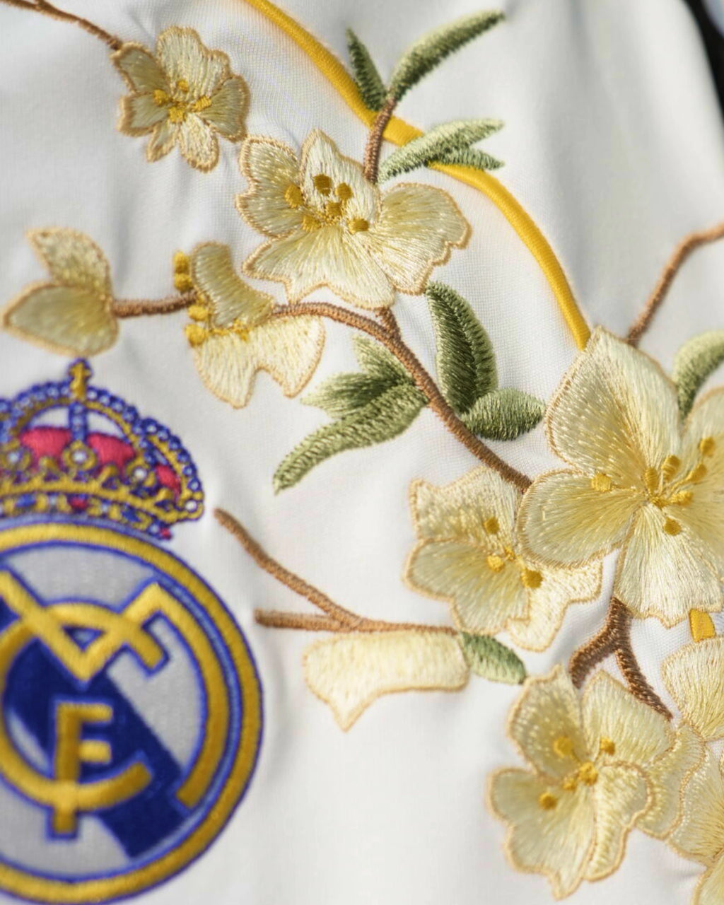 Close-Up Macro Shot Focus On The Left Chest Of The Real Madrid Home Kit Revisited, Highlighting The Dense Haptic 3D Thread Quality Of Pale Gold Flowers Encasing The Crowned Crest.