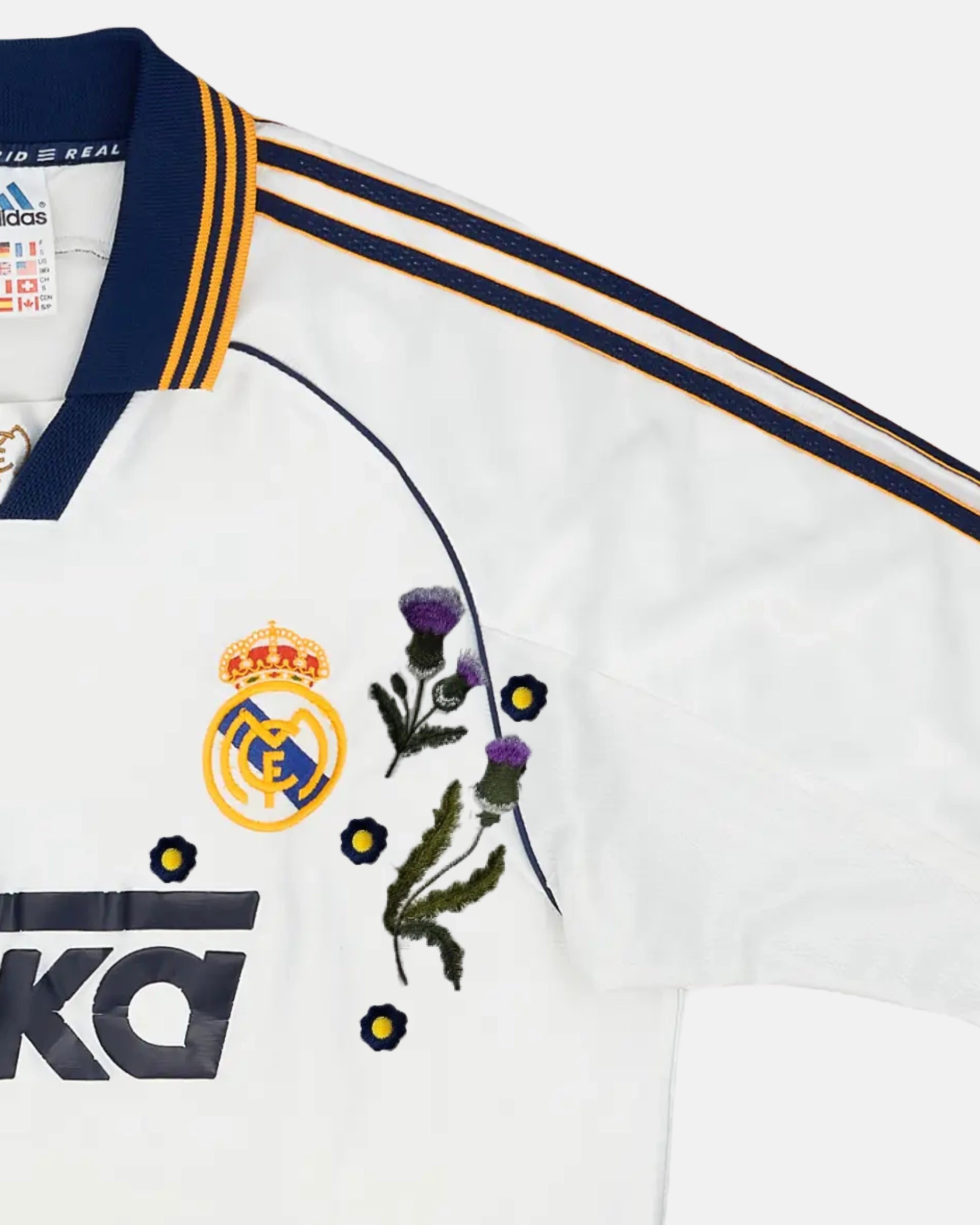 Close-Up Detail Of The Real Madrid Vintage Kit Revisited Right Chest, Illustrating Dense Purple and Green Flower Stitches Next To The Crowned Royal Crest.
