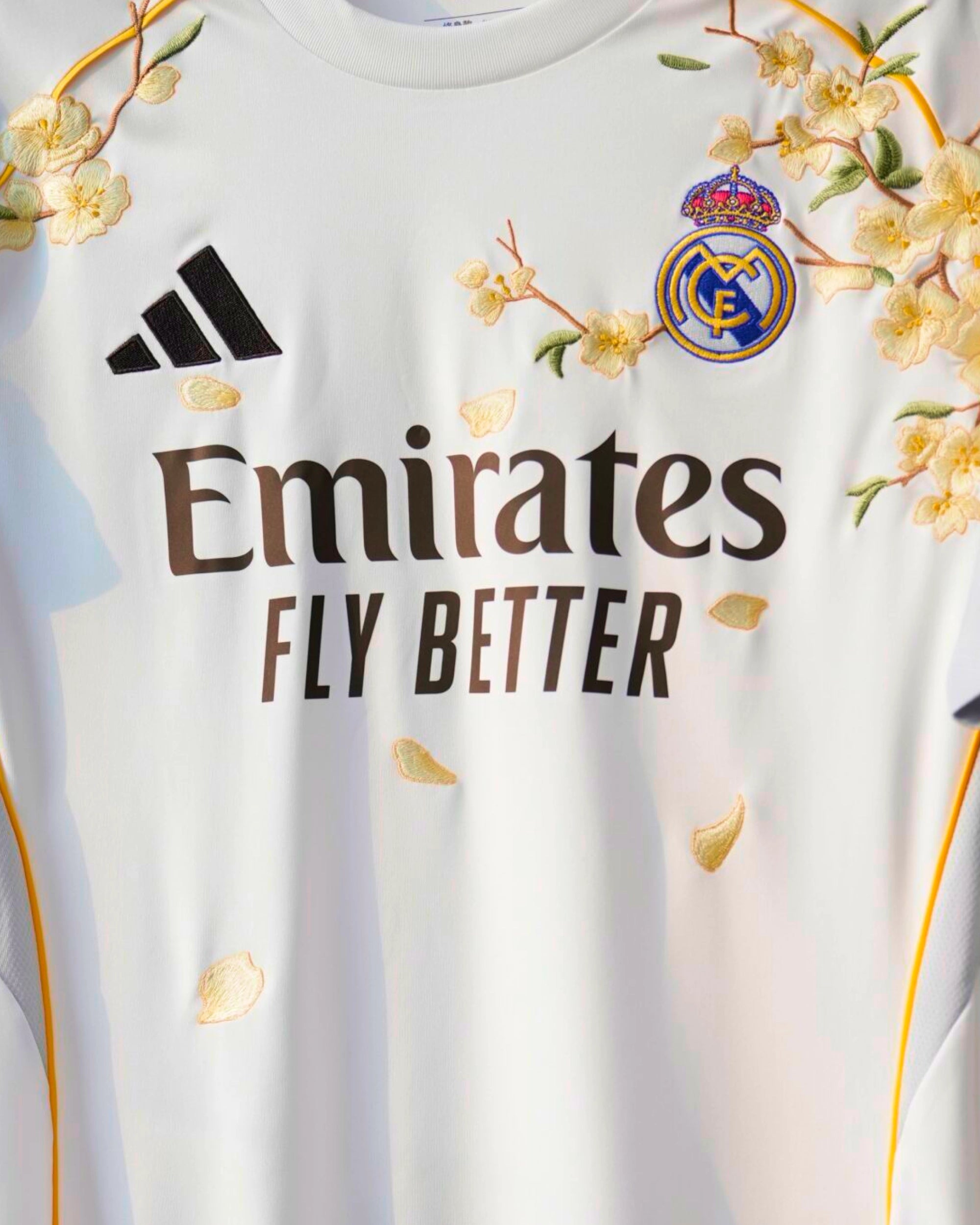 Medium Chest Shot Detail Of The Real Madrid White Jersey Reimagined, Showcasing Elegant Golden-Yellow Flower Embroidery Winding Above The Dark Corporate Sponsor Text.